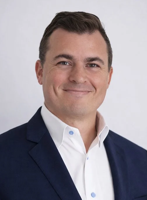 A professional headshot of a smiling man in a dark suit and white shirt against a plain background.