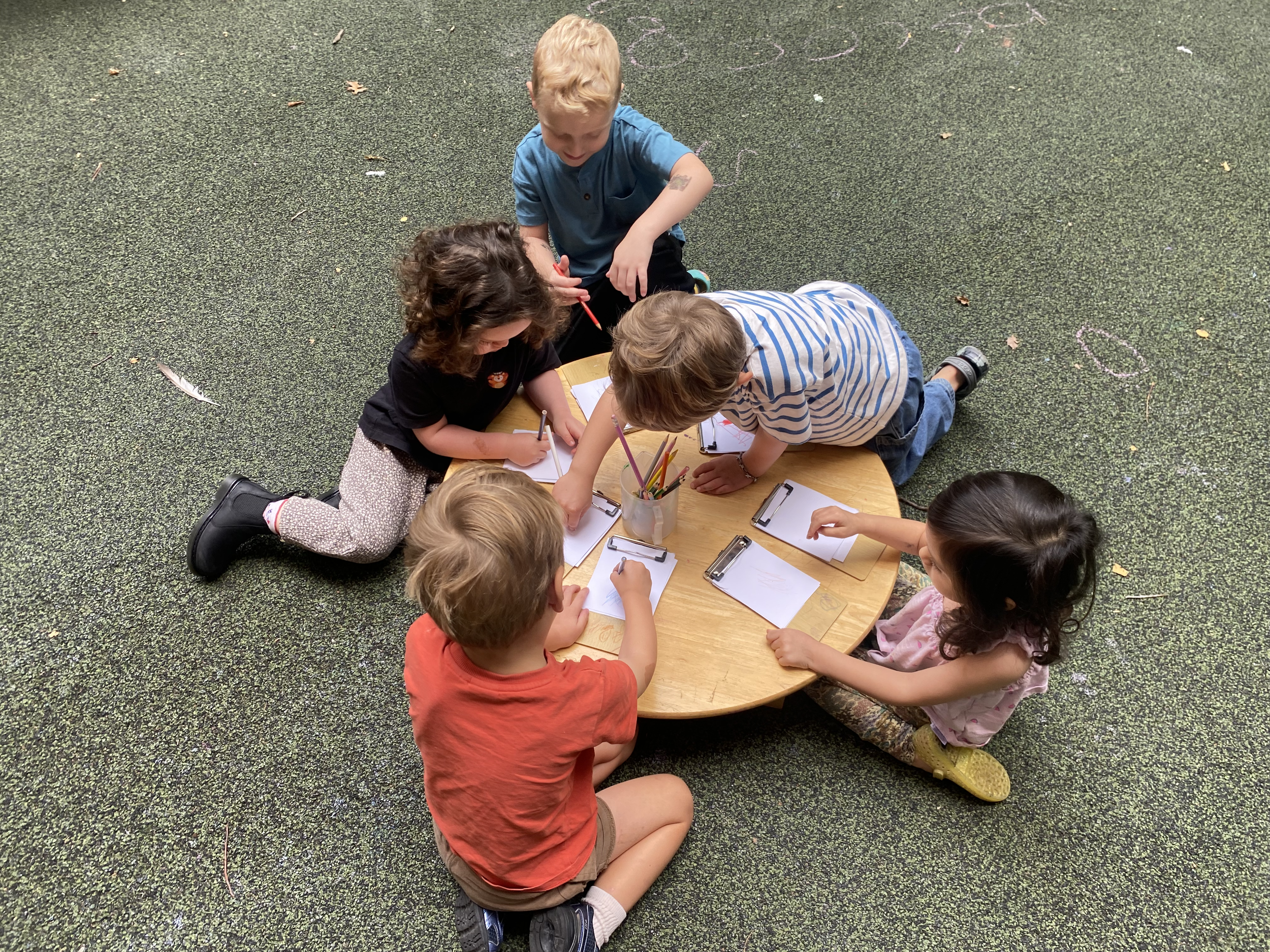 Six young children gathered around a small wooden table on the floor, drawing and coloring on clipboards with various pens and markers.