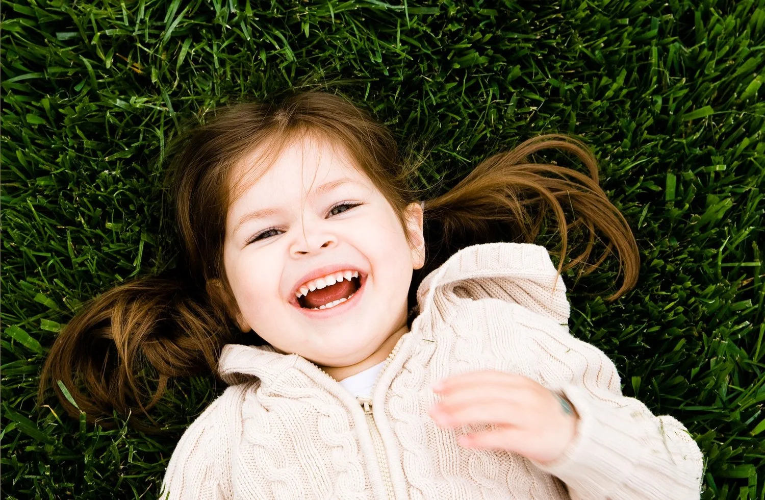 A young girl with brown hair lying on green grass, smiling and laughing with her mouth open.