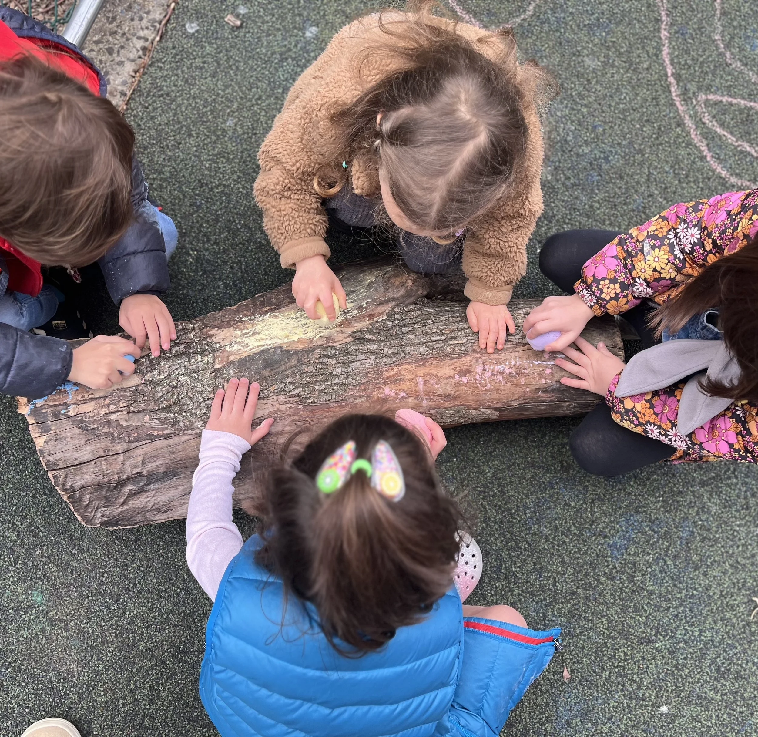 Children drawing on a large piece of logs with chalk on a textured ground.