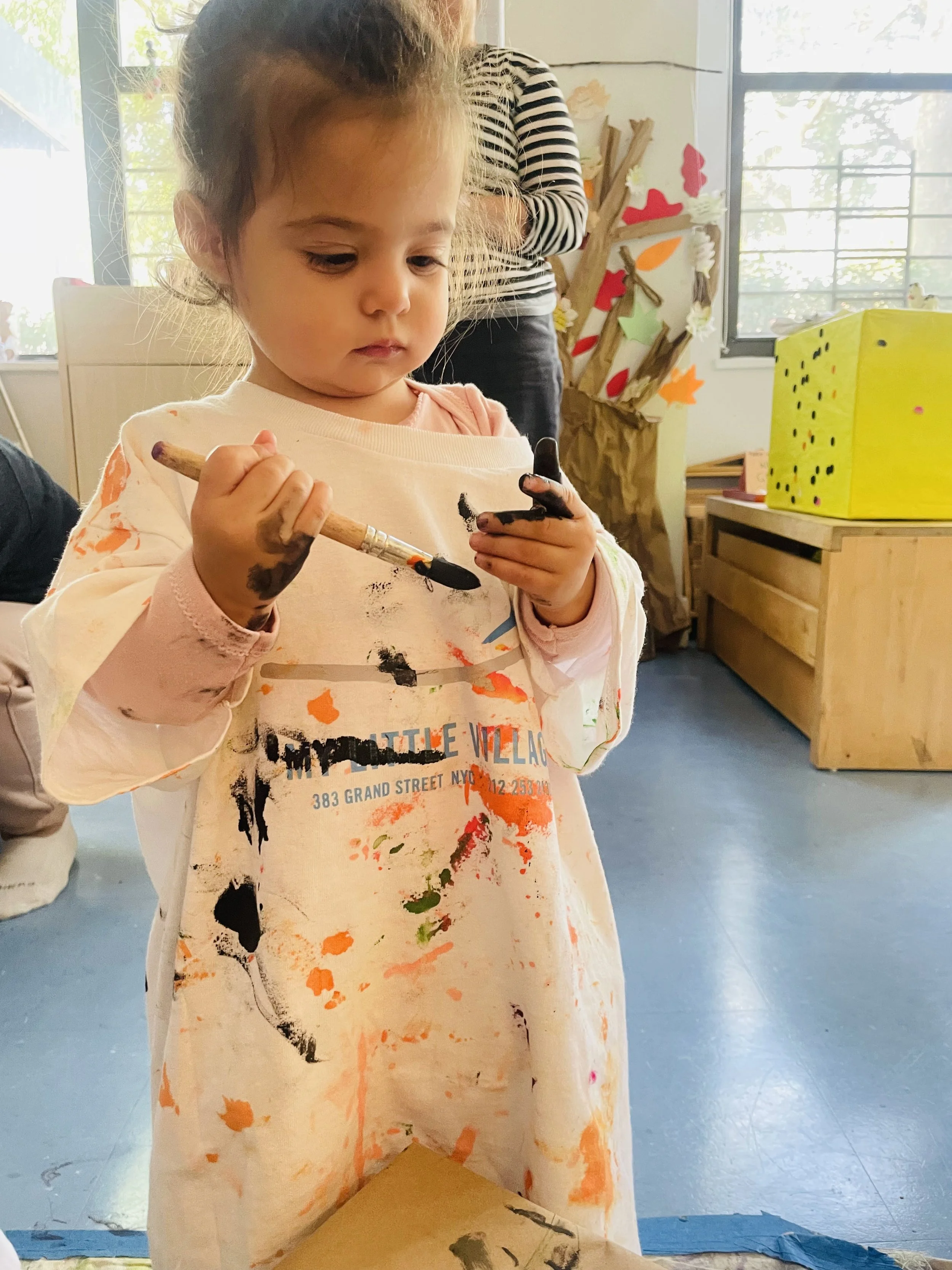 Young girl with paint on her hands and clothes holding a paintbrush and a small object, engaging in a painting activity in an art classroom.