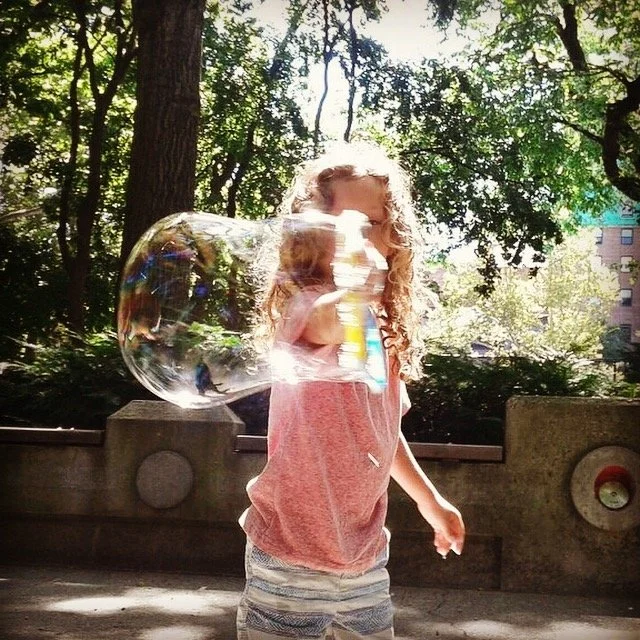 A young girl with curly hair wearing a pink shirt and striped shorts playing with a large bubble outdoors in a park during daytime, with trees and buildings in the background.