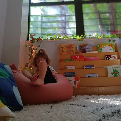 A young child with curly hair sitting on a pink bean bag chair in a cozy room with a white shag rug, next to a bookshelf filled with children's books, and a window with green foliage outside.
