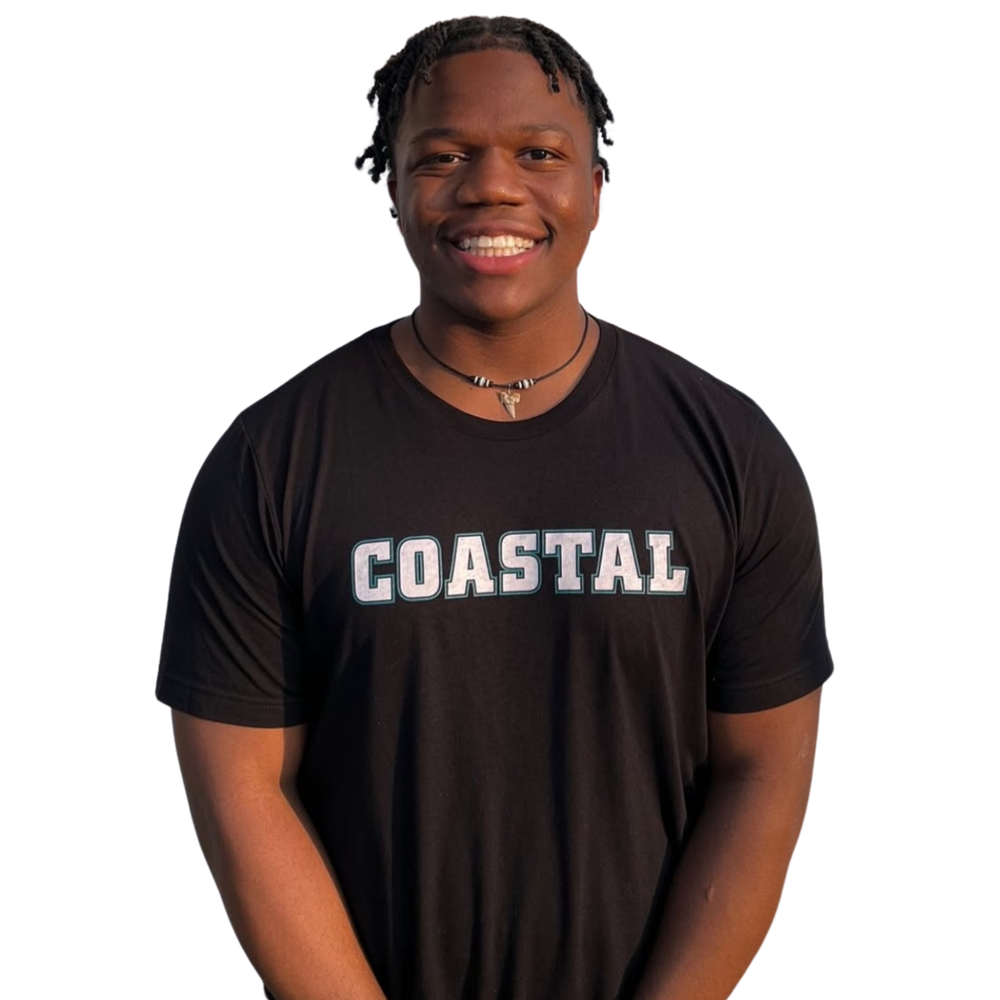 Smiling young man with short dreadlocks, wearing a black t-shirt with the word 'COASTAL' printed on it, and a necklace with a shell pendant against a dark background.