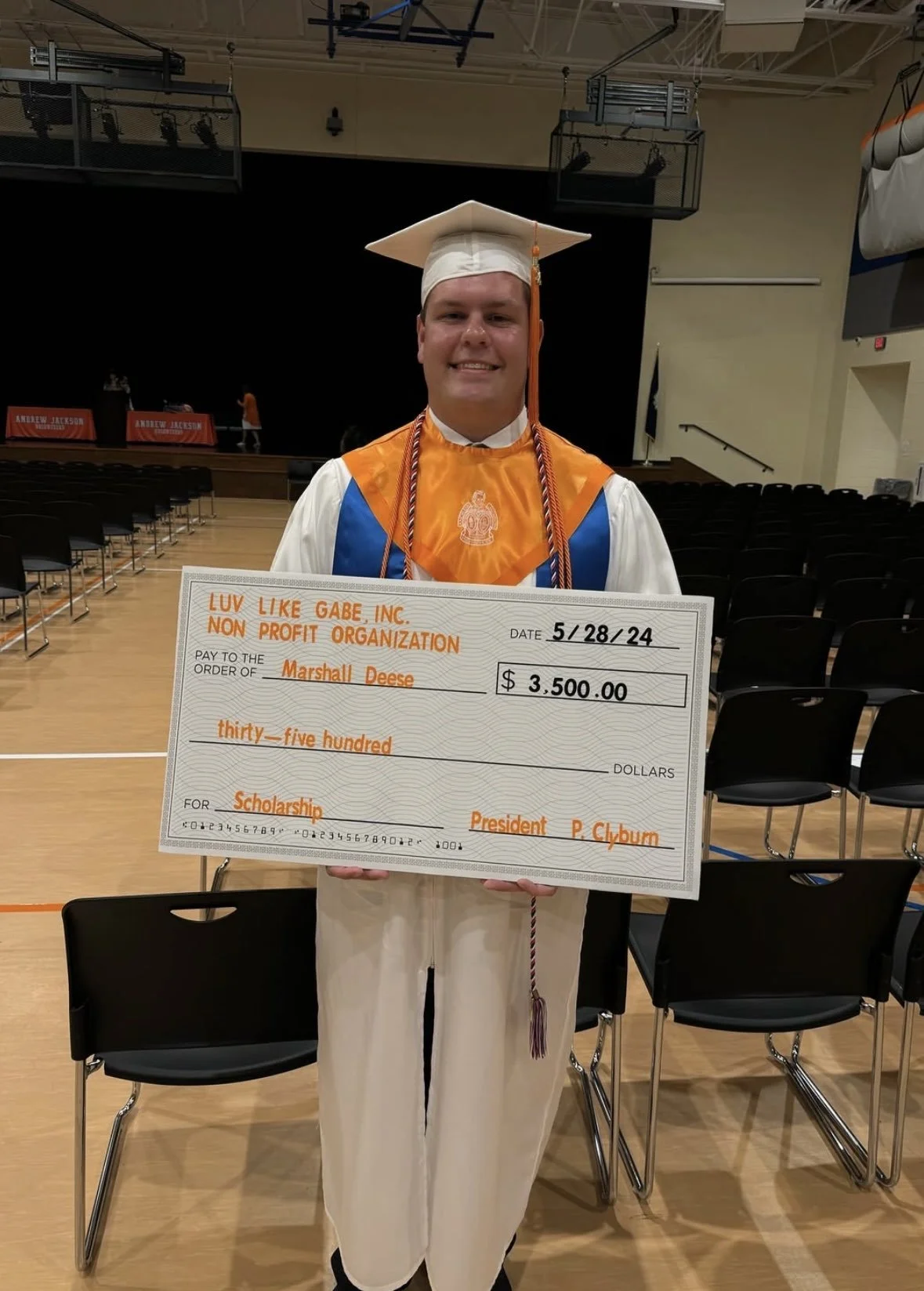 A young man in a graduation gown and cap standing in a gymnasium, holding an oversized check for $3,500 made out to Marshall Deese for a scholarship, with the date 5/28/24, and a smile on his face.