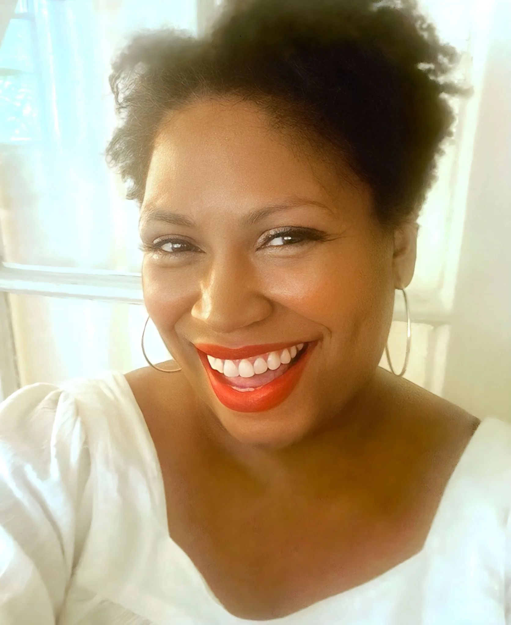 Close-up of a woman smiling with red lipstick, wearing hoop earrings and a white top, standing in front of a window with sheer curtains.