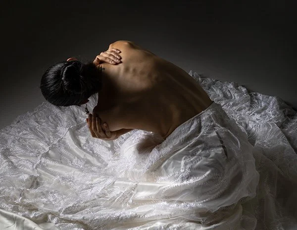A topless woman with braided hair sitting on a bed with white satin sheets, holding her neck and shoulder with her head bowed.