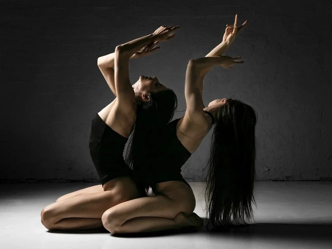Two women practicing yoga together in a dark room, sitting on their heels with their backs arched, hands raised, and hair hanging downward.