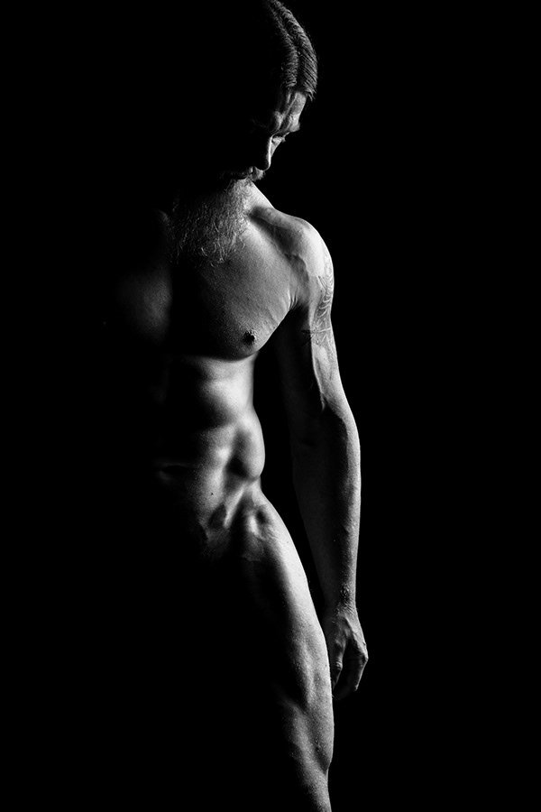 A black and white photo of a shirtless man with long hair and a beard, standing in profile with his head bowed and eyes closed, against a dark background.