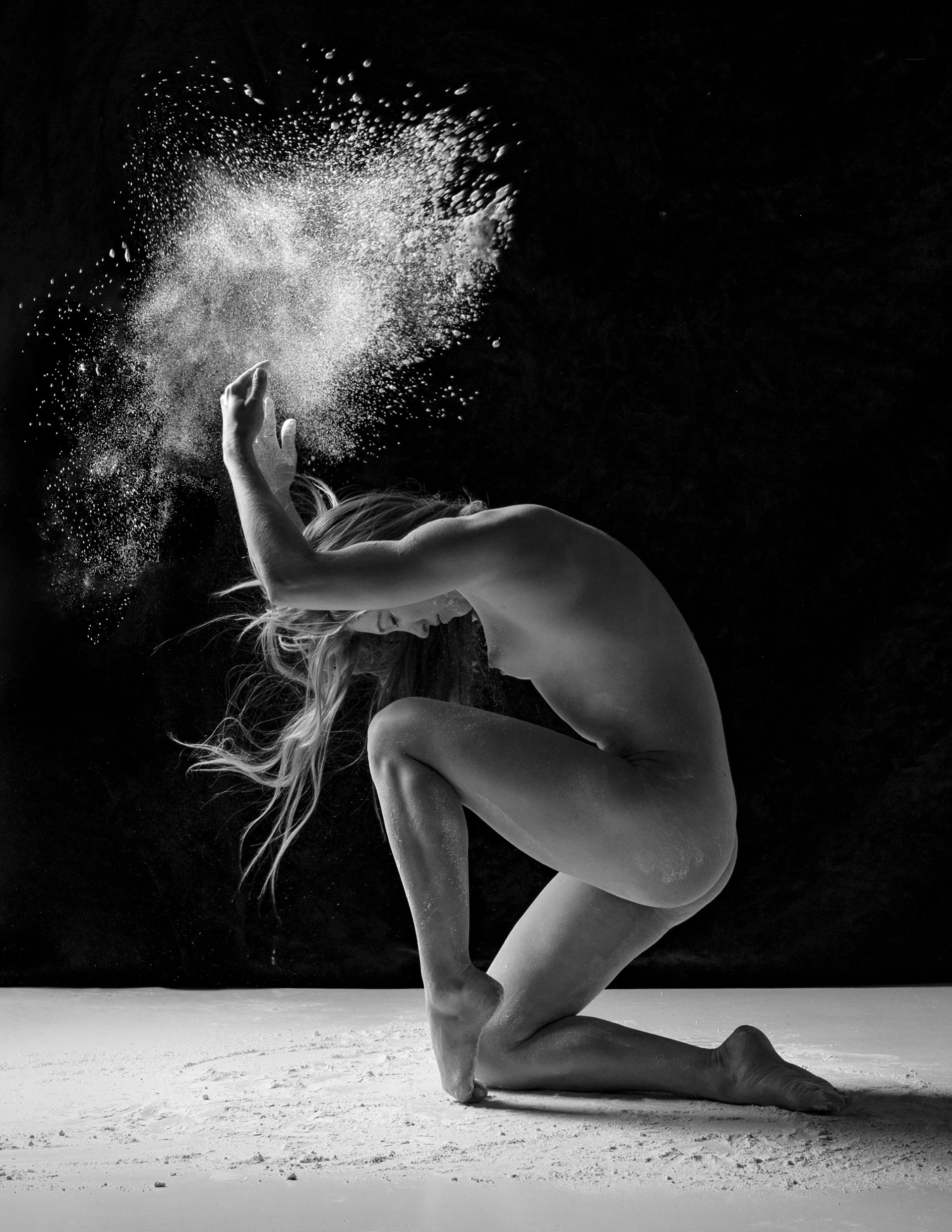 A nude woman kneeling on the ground, throwing a cloud of powder into the air, captured in black and white.