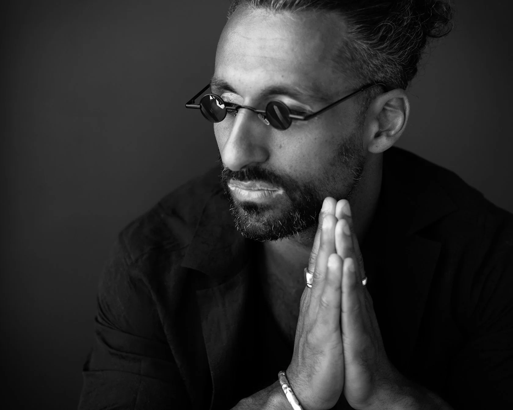 A black-and-white photo of a man with glasses, a beard, and styled hair, sporting rings and a bracelet, with hands pressed together in a prayer-like pose, looking down thoughtfully.