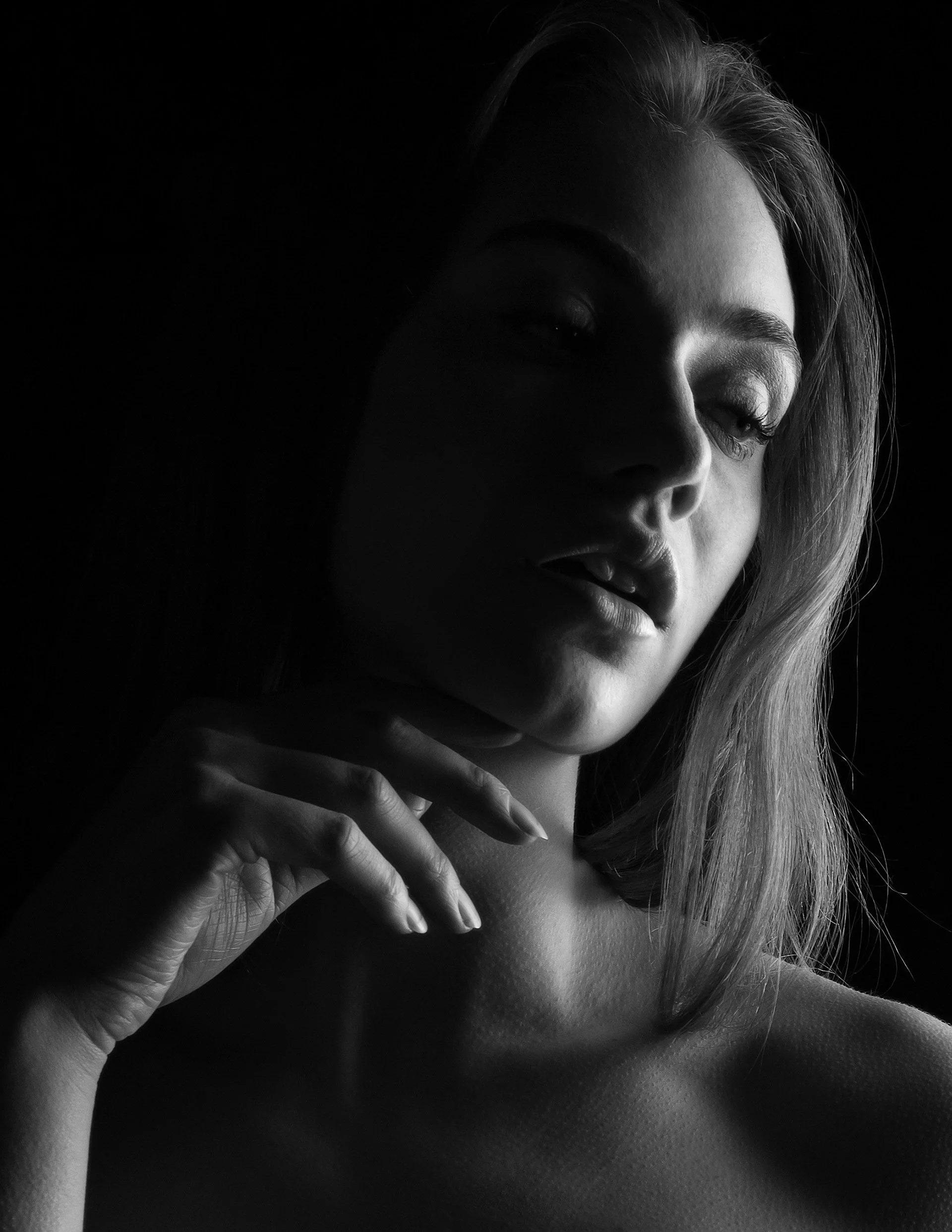 A black and white portrait of a woman with light skin, partially illuminated, with her hand gently touching her neck and shoulder.