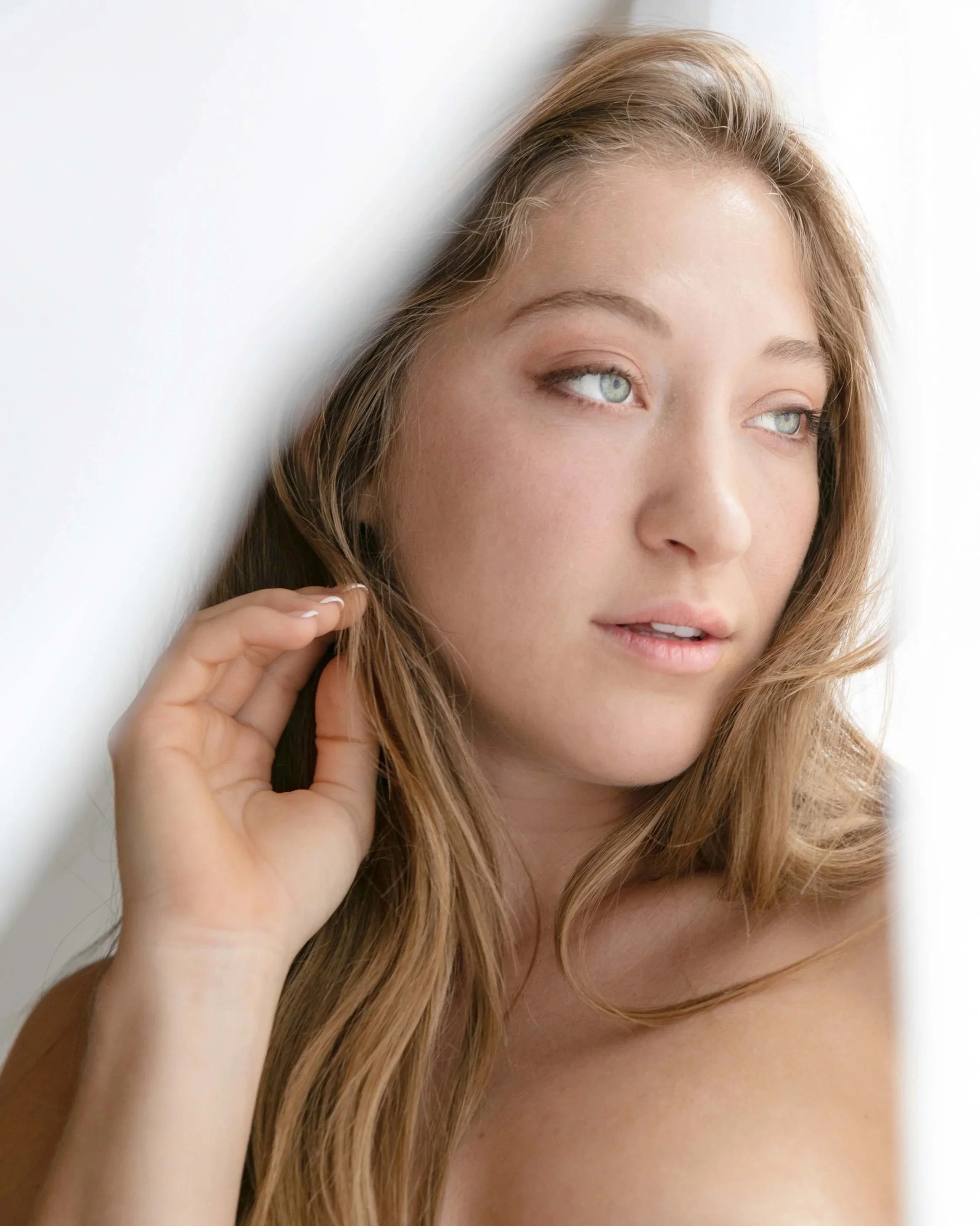A young woman with long blonde hair and light skin looking to the right, touching her hair with her left hand, against a soft white background.