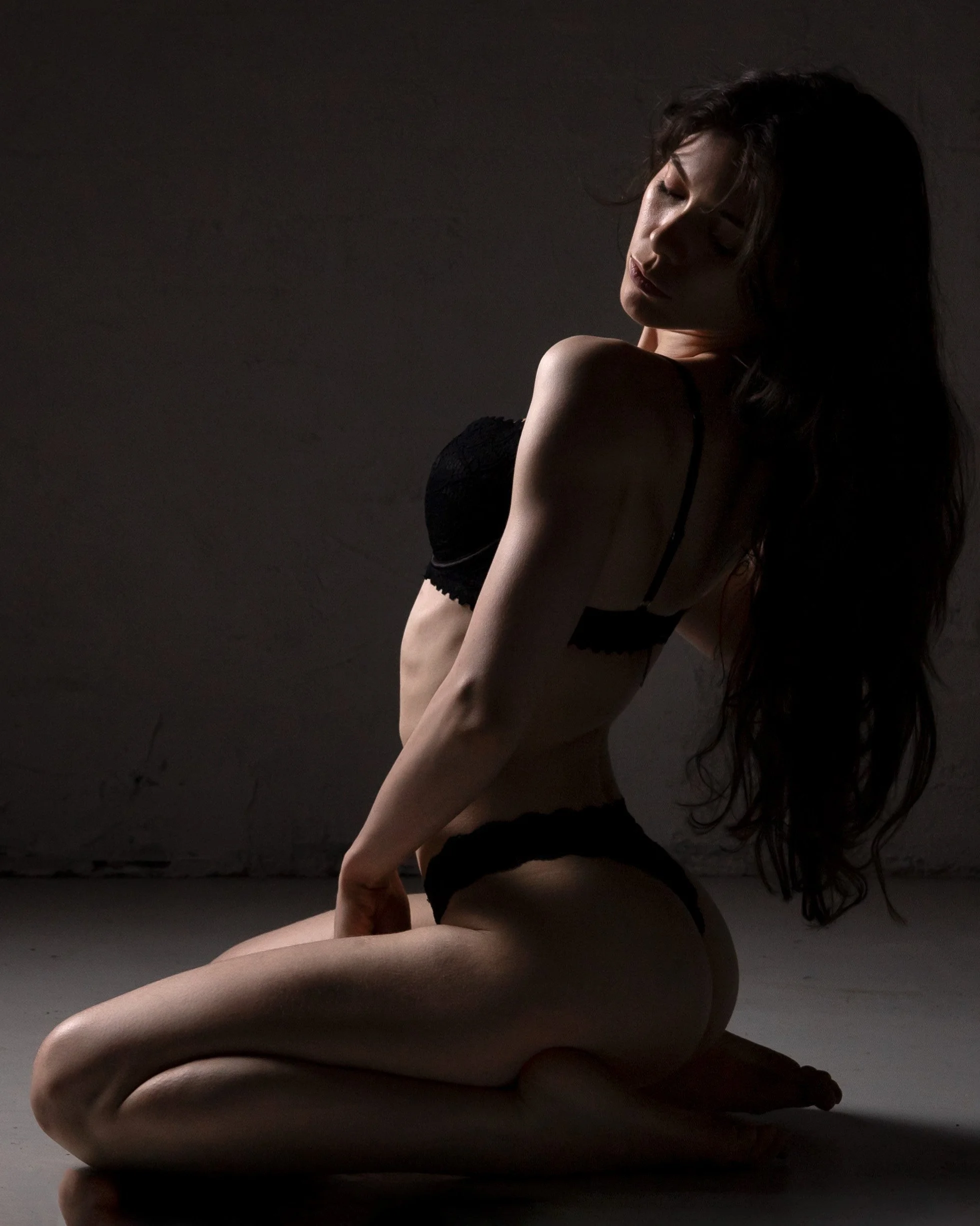 A woman kneeling on the ground in dark lingerie, posing with one arm resting on her thigh, in a low-lit setting with a textured wall background.