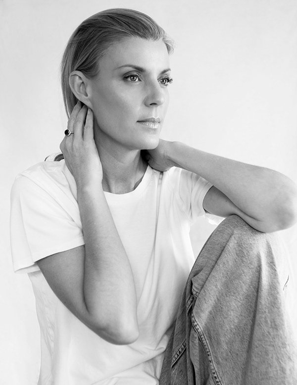 Black and white portrait of a woman wearing a white t-shirt and jeans, sitting with one knee up, touching her neck and ear, with a calm and contemplative expression.