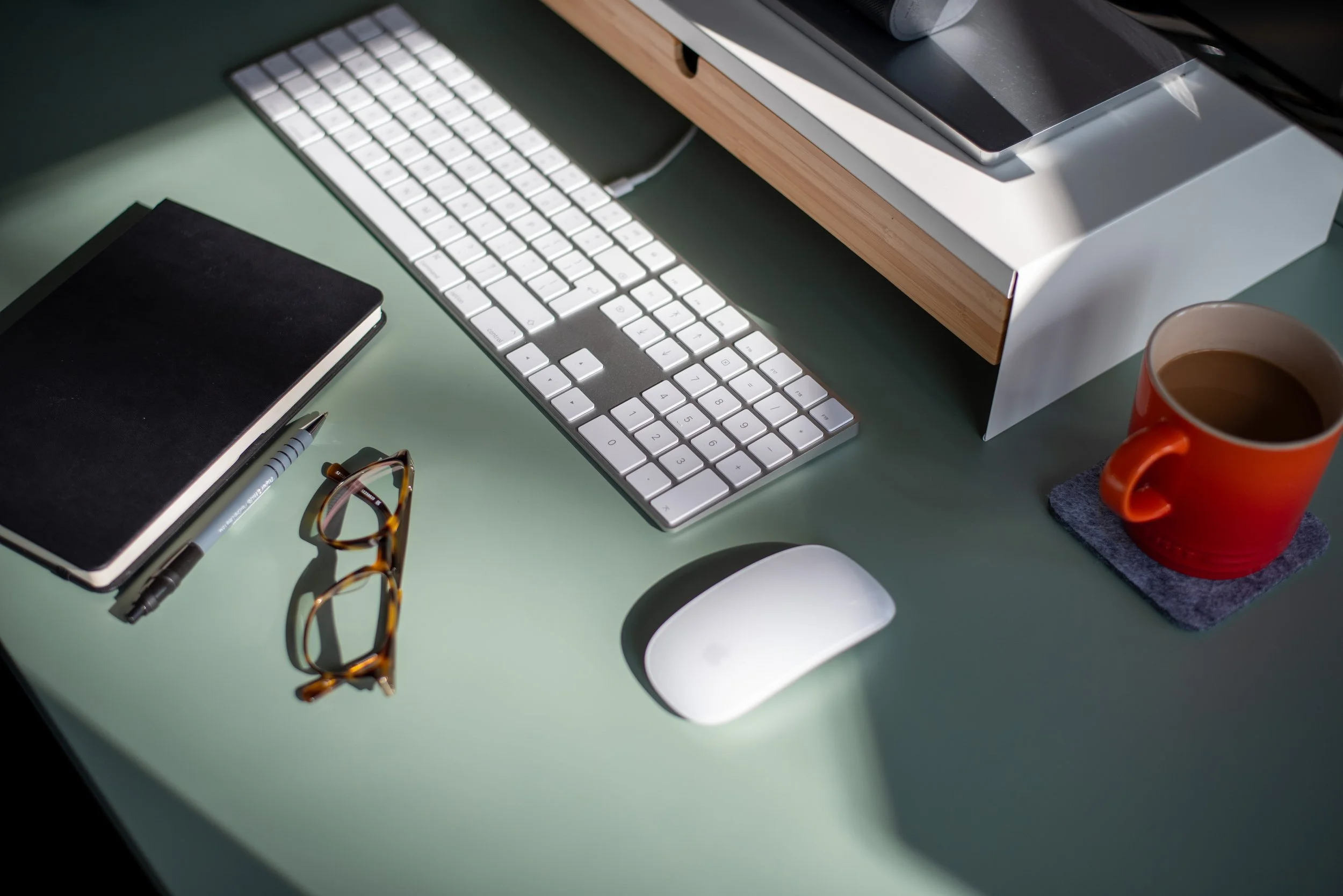 Work desk with a keyboard, mouse, notebook, pen, glasses, and a cup of coffee on a coaster.