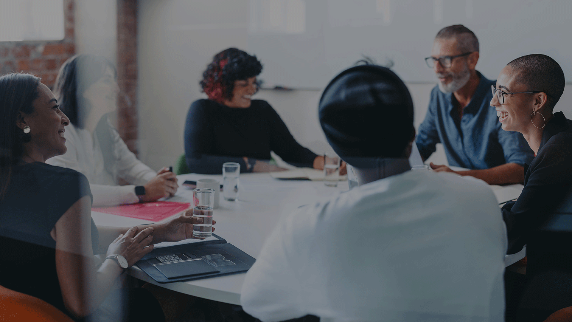 A diverse group of six people, four women and two men, sitting around a conference table in a modern office, smiling, engaged in conversation, with laptops, tablets, and glasses of water.