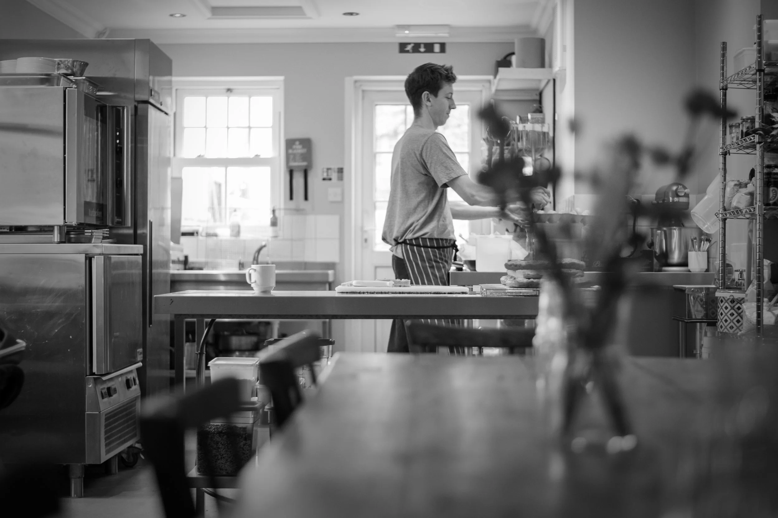 A person in a kitchen wearing an apron preparing food on a countertop, with shelves and appliances visible in the background.