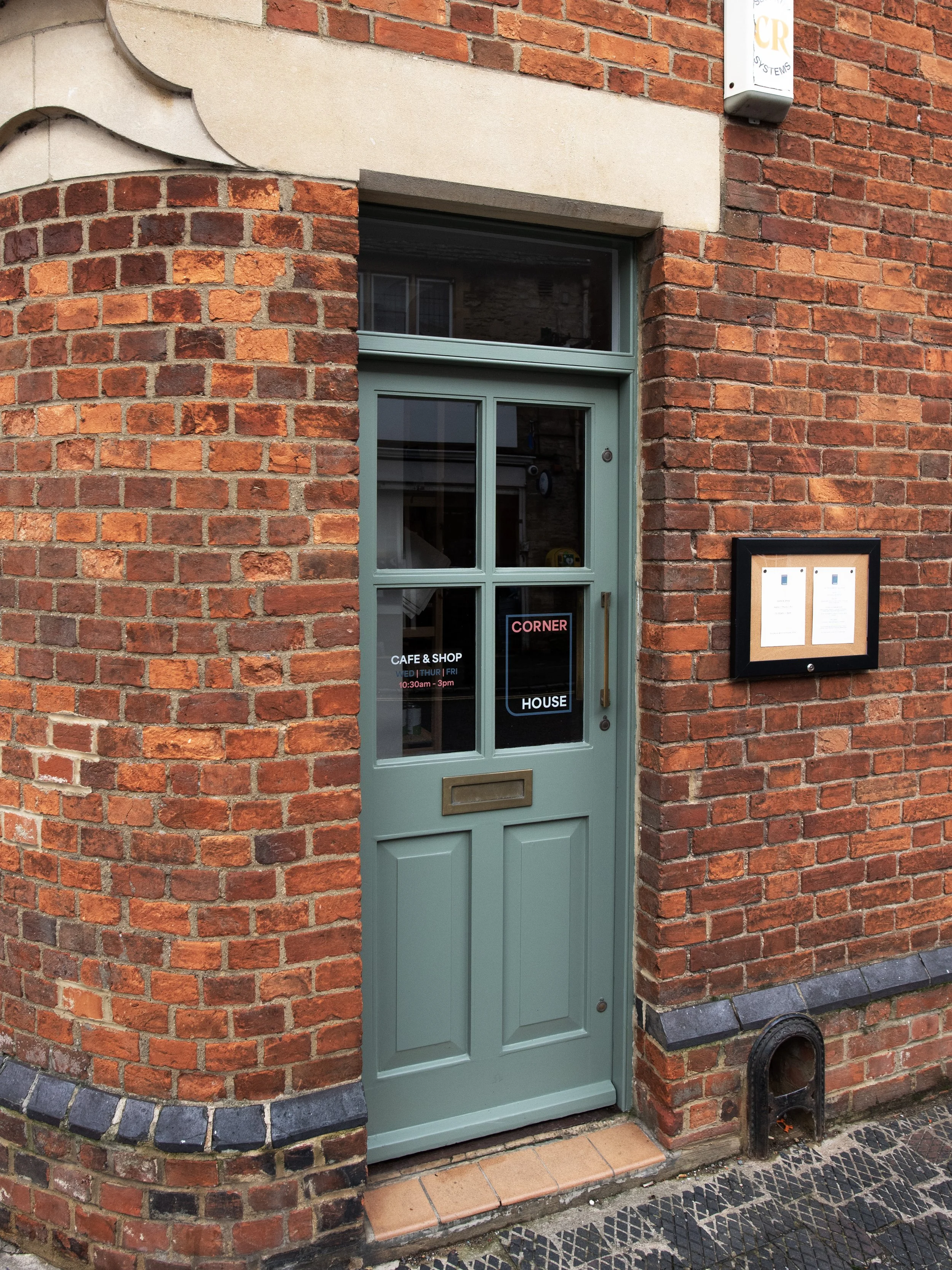 Green door of a cafe and shop named 'Corner House' with a black-framed menu display on a red brick building.