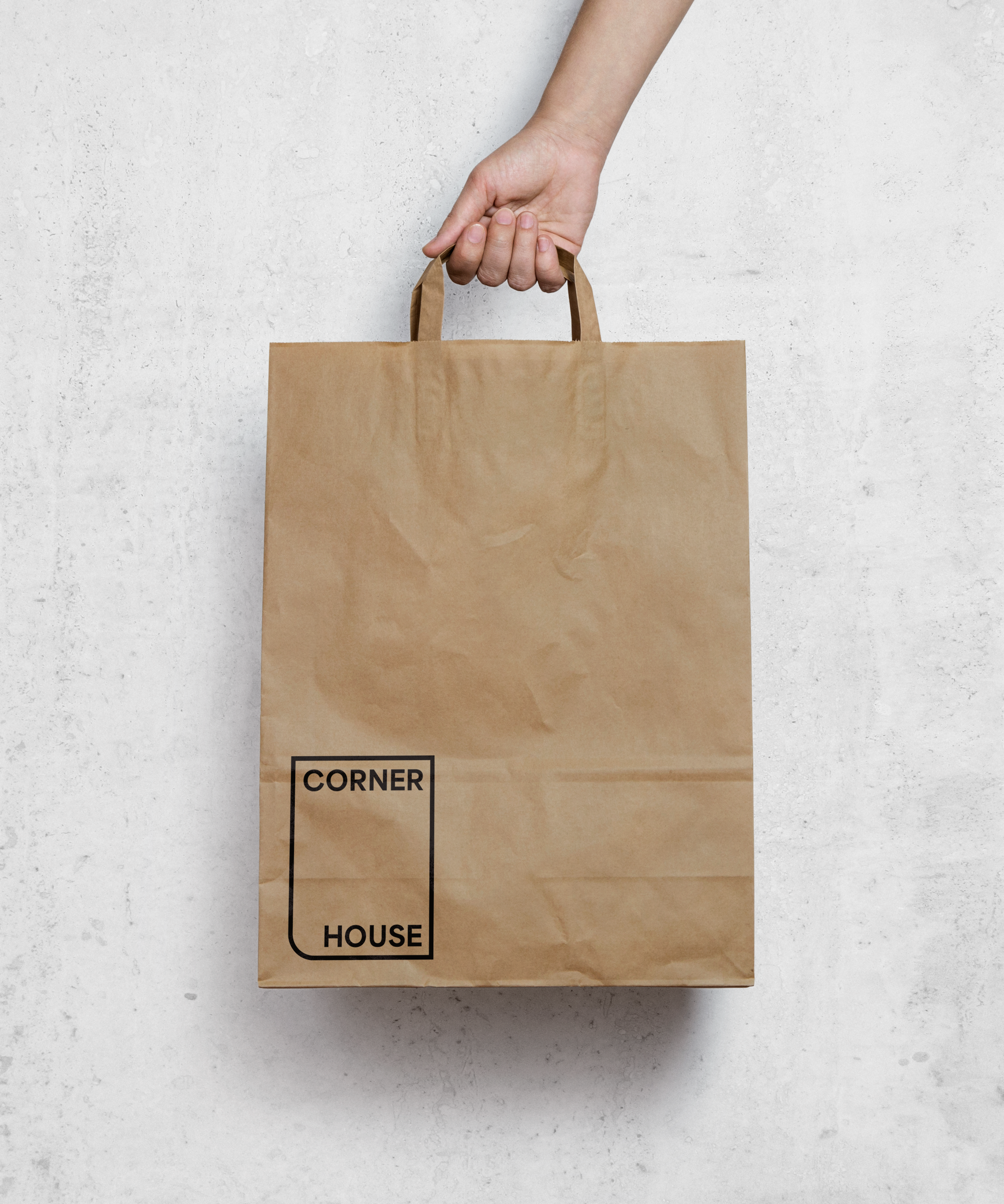 A hand holding a brown paper bag against a white textured wall. The bag has the text 'CORNER HOUSE' printed in black in a rectangular outline.