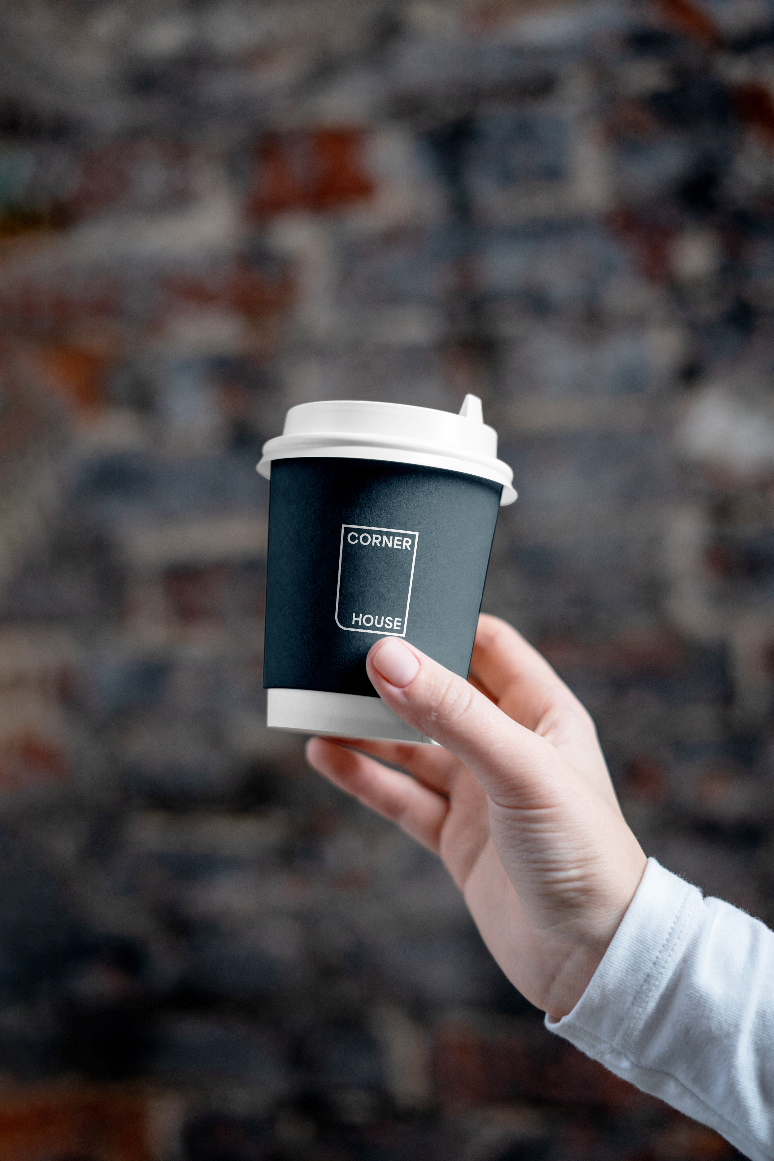 A hand holding a white disposable coffee cup with a black sleeve that has a minimalist logo reading 'CORNER HOUSE' against a blurred brick wall background.