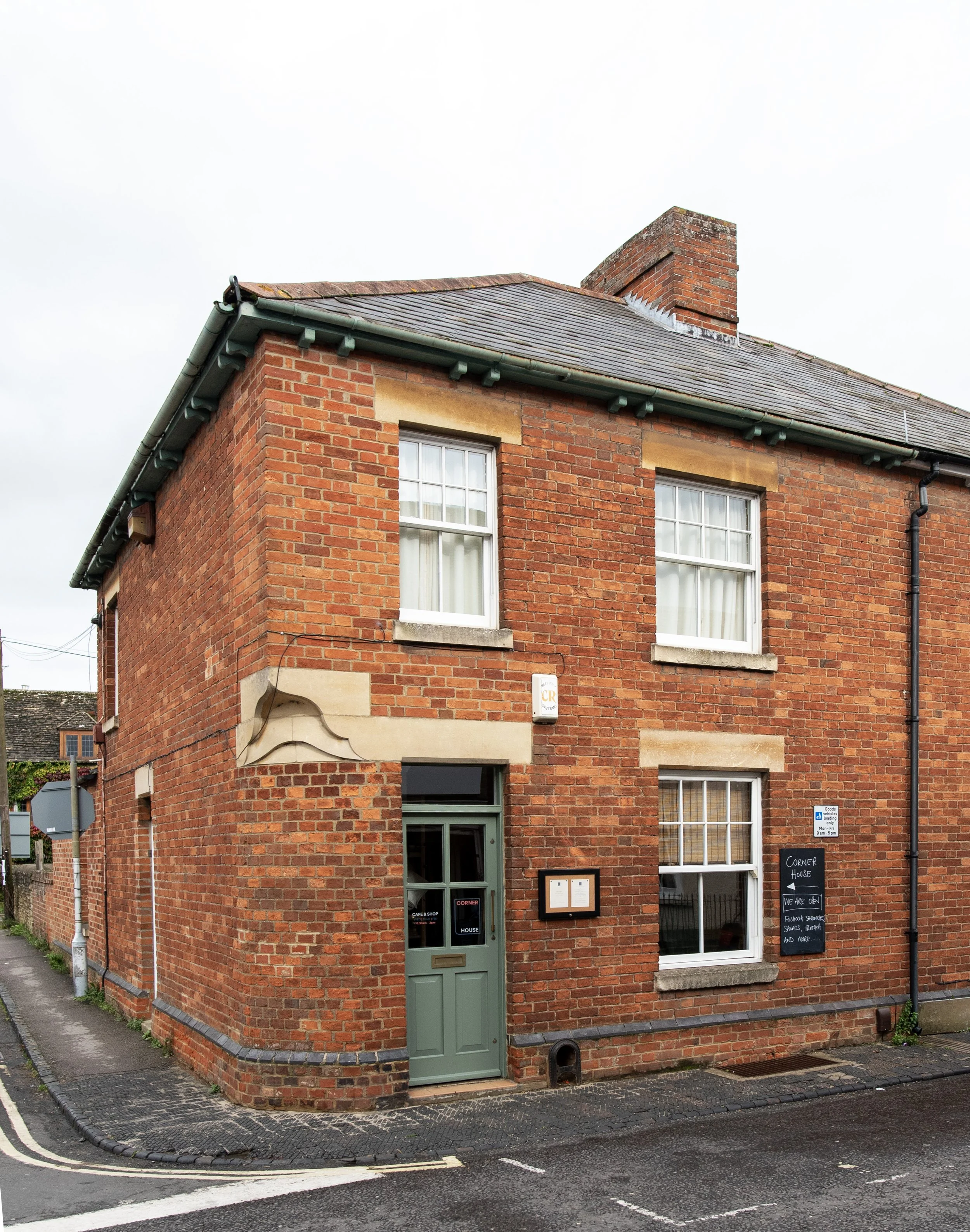 A two-story brick building with a green door and three white double-hung sash windows, located on a street corner with a blackboard sign and parking signs outside.