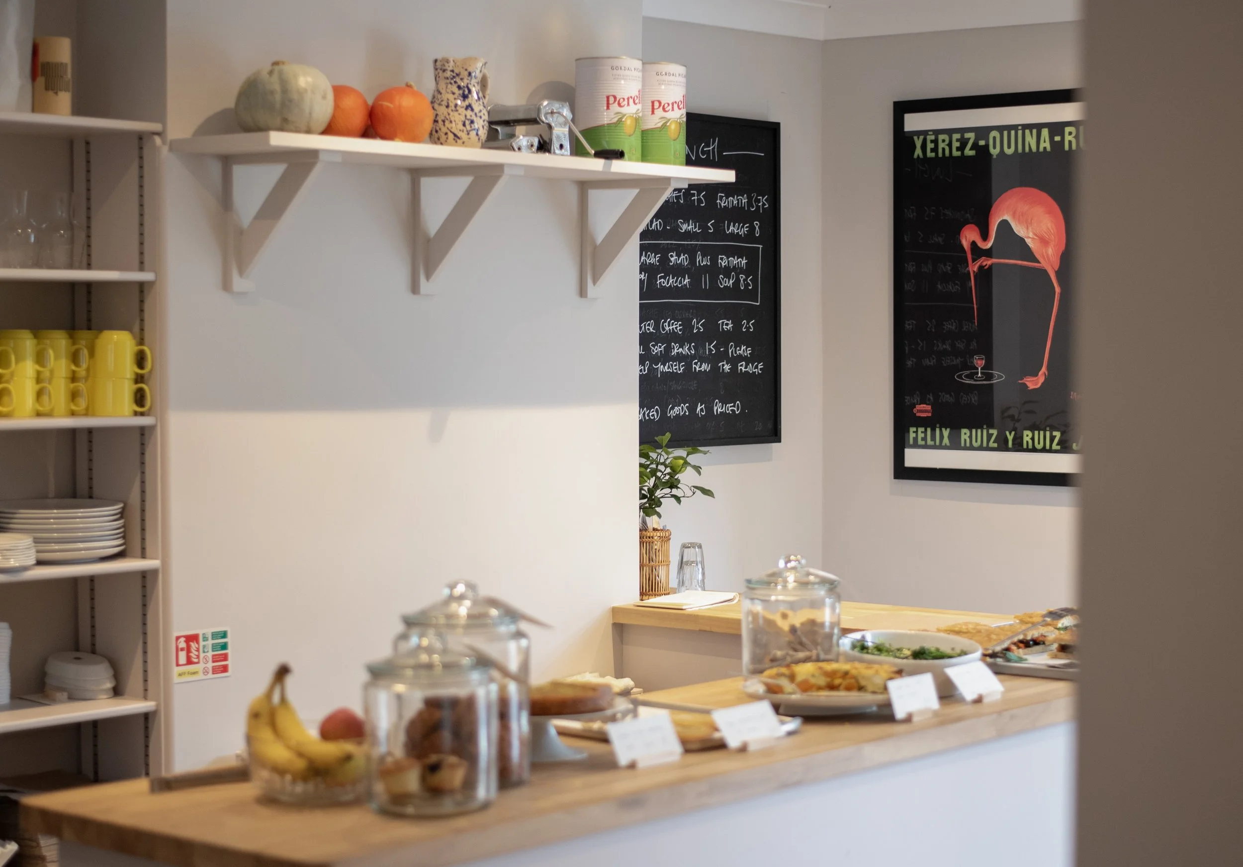 A cafe counter with food and drinks, including bananas, fruit, and pastries in glass jars, with a menu and flamingo poster on the wall.