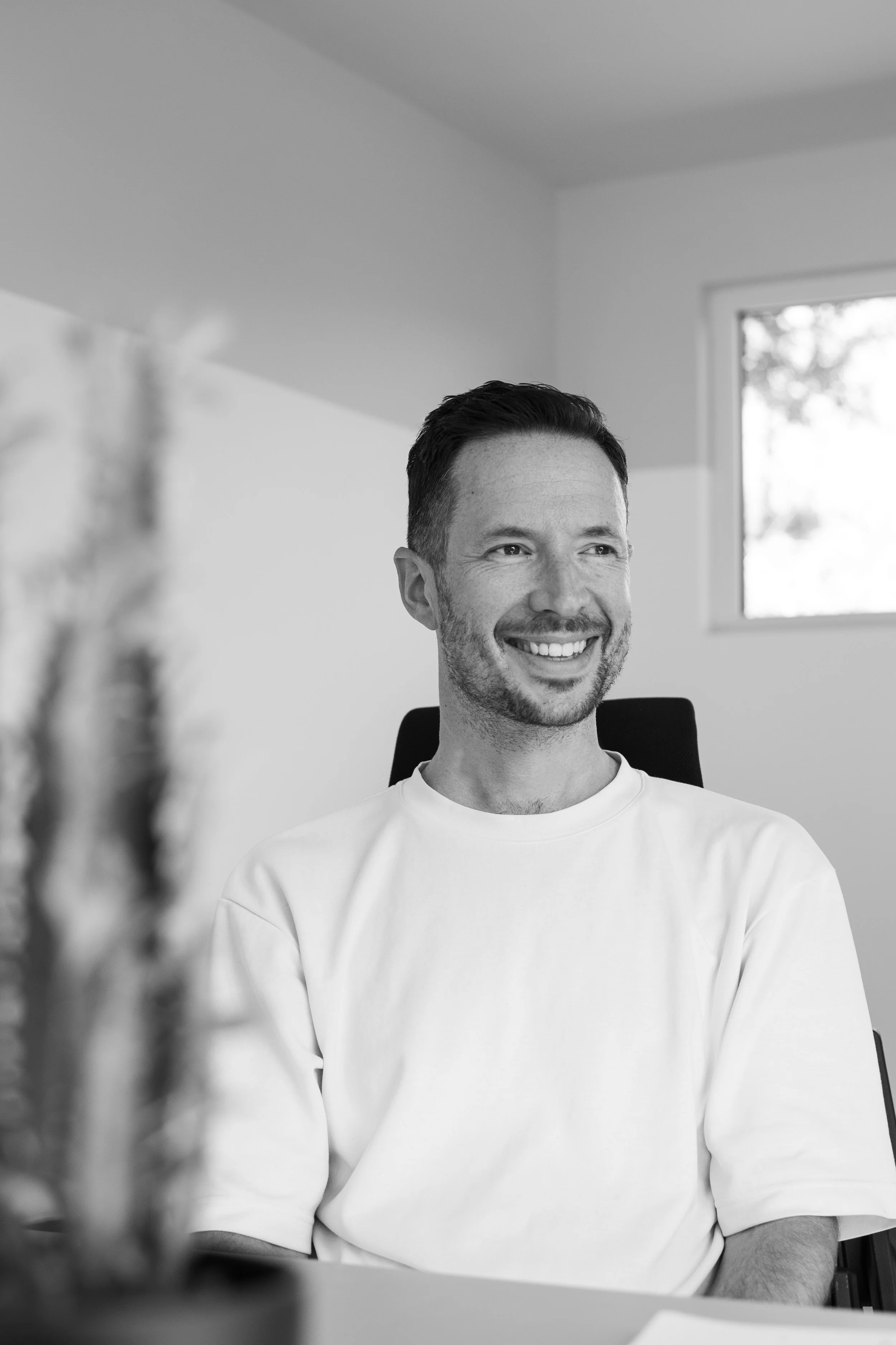 A smiling man with short hair and a beard sitting in a chair in a well-lit room with a window in the background.