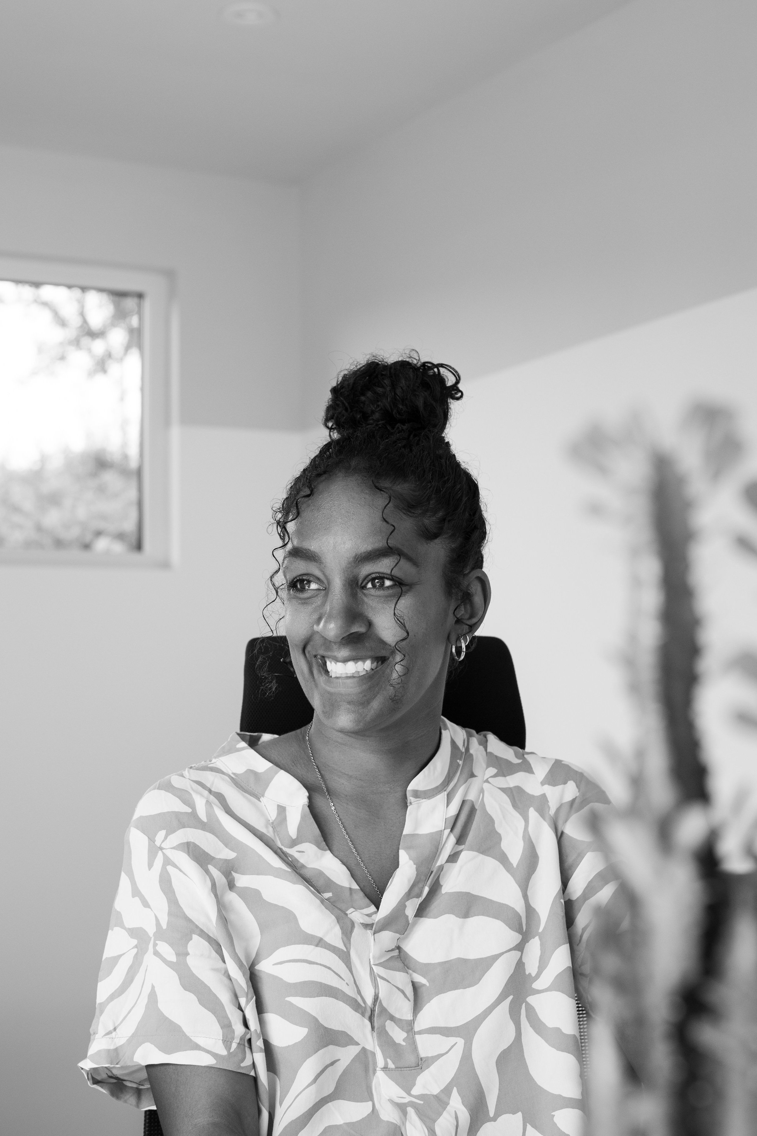 Black and white photo of a woman with a bun hairstyle, smiling, wearing a patterned blouse, sitting in a room with a window and framed wall art in the background.