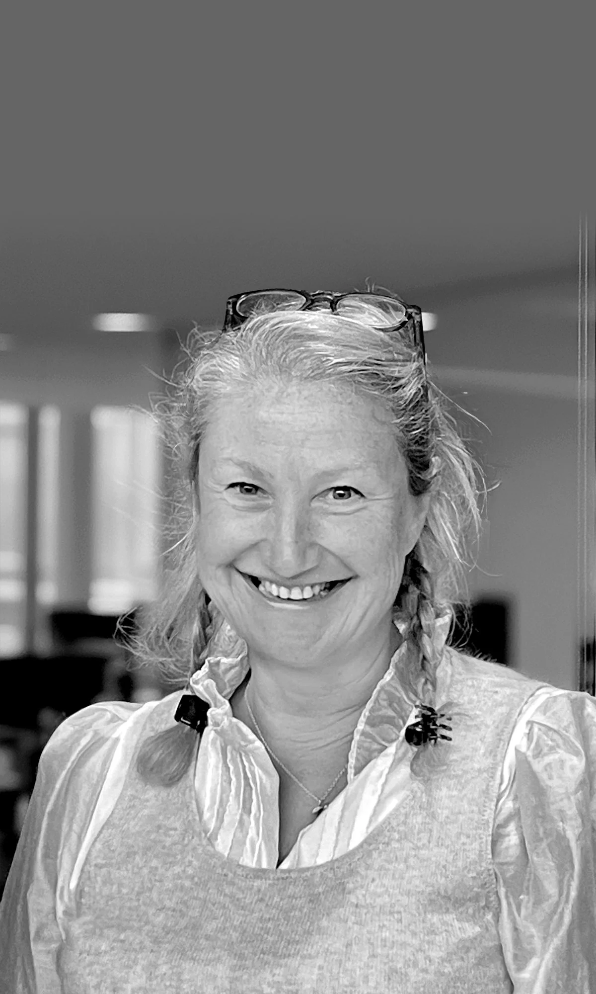 A smiling woman with glasses resting on her head, wearing a striped shirt under a light-colored vest, standing indoors.