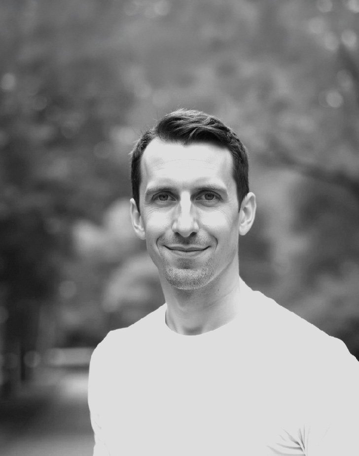 Black and white portrait of a man with short, dark hair, smiling, wearing a white shirt, outdoors with blurred trees in the background.