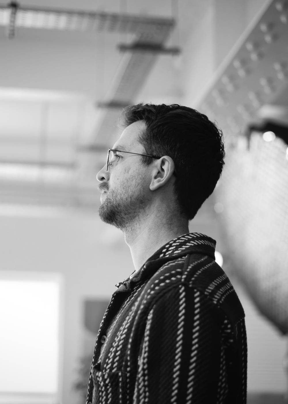 Profile of a young man with glasses and dark hair, standing indoors with a blurred background