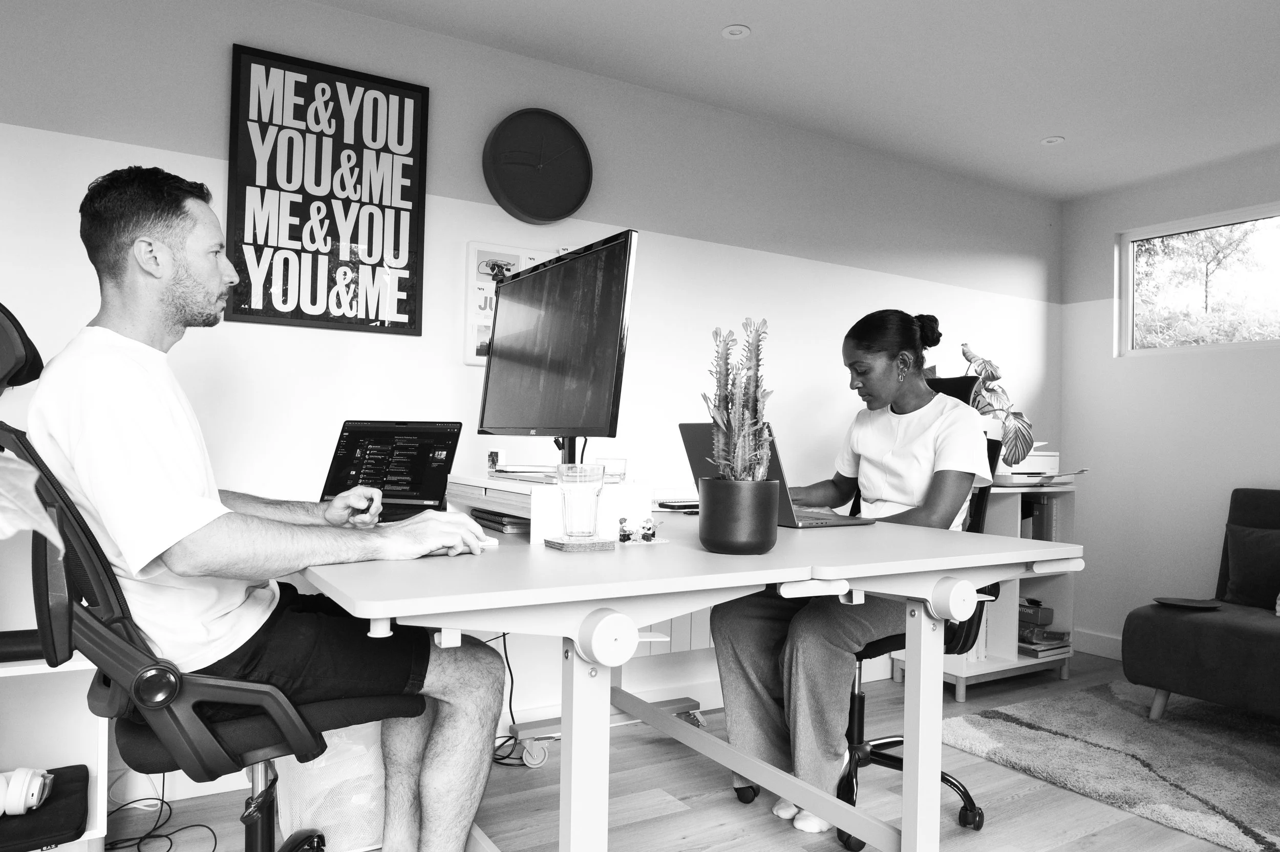 Two people working at a desk in a modern office, with multiple monitors and plants, with a framed poster reading 'ME & YOU' on the wall.