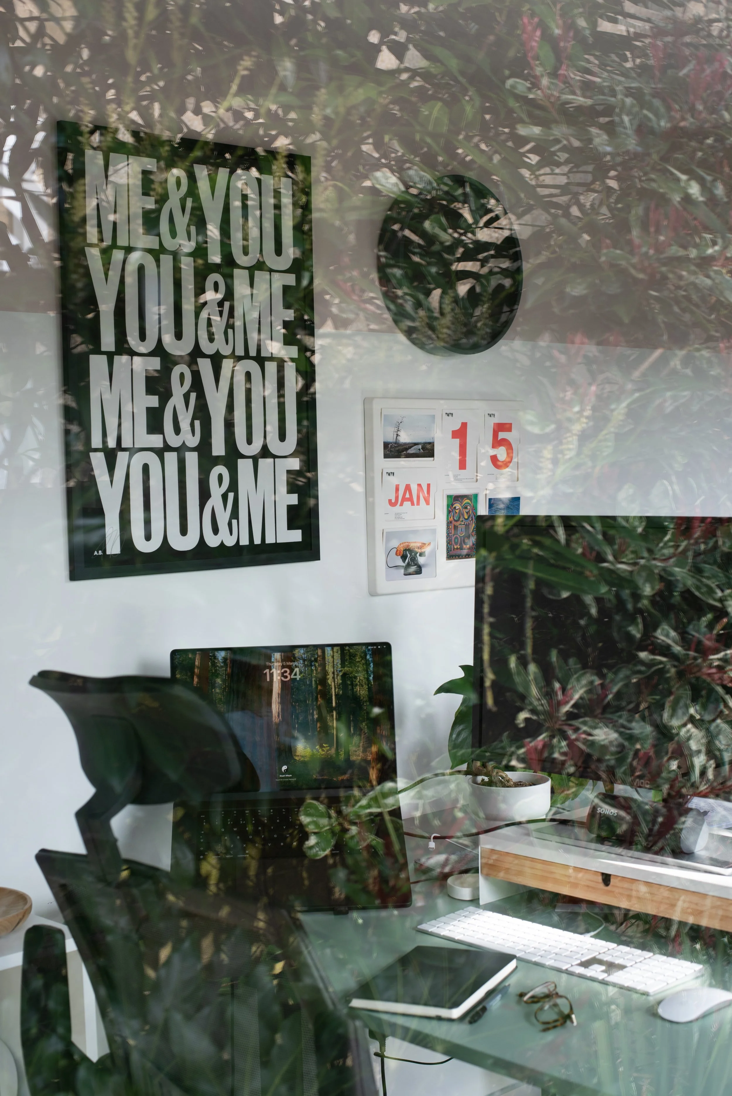 A home office seen through a glass window, with plants and decor including a black and white poster with repeated text 'ME & YOU & ME & YOU' and a calendar displaying January 15. On the desk, there is a monitor, a keyboard, a tablet, and a pair of glasses. Reflections of trees and outdoors are visible on the glass.