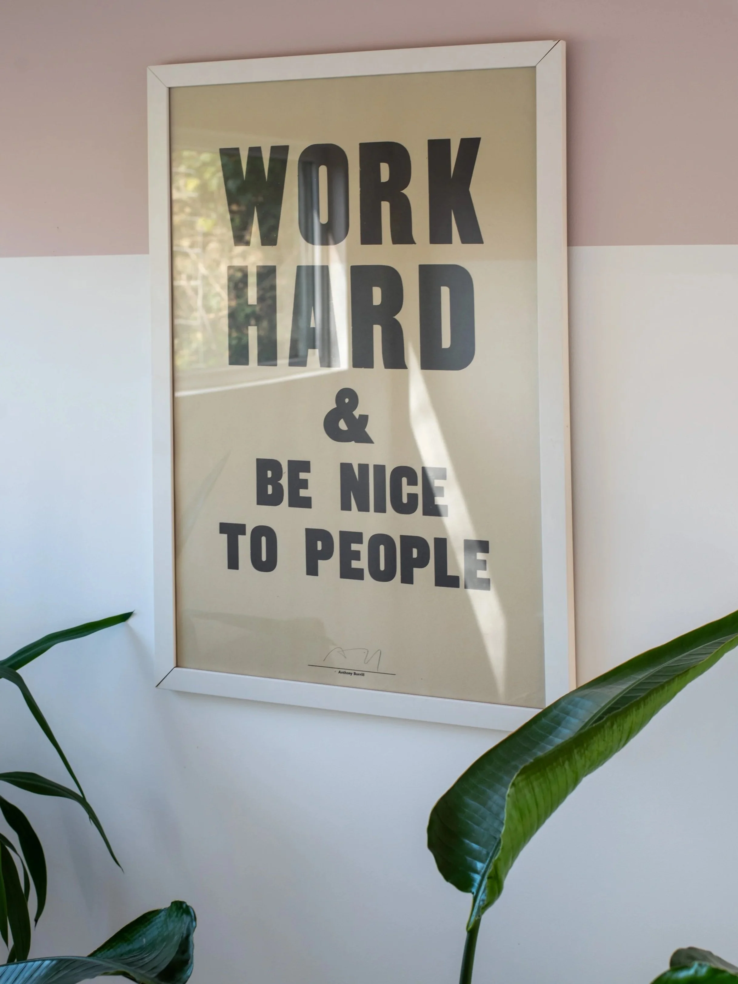 A framed poster on a wall with the text 'Work Hard & Be Nice to People' in bold black letters. The wall background is painted in light pink and white, with some green plants in the foreground.
