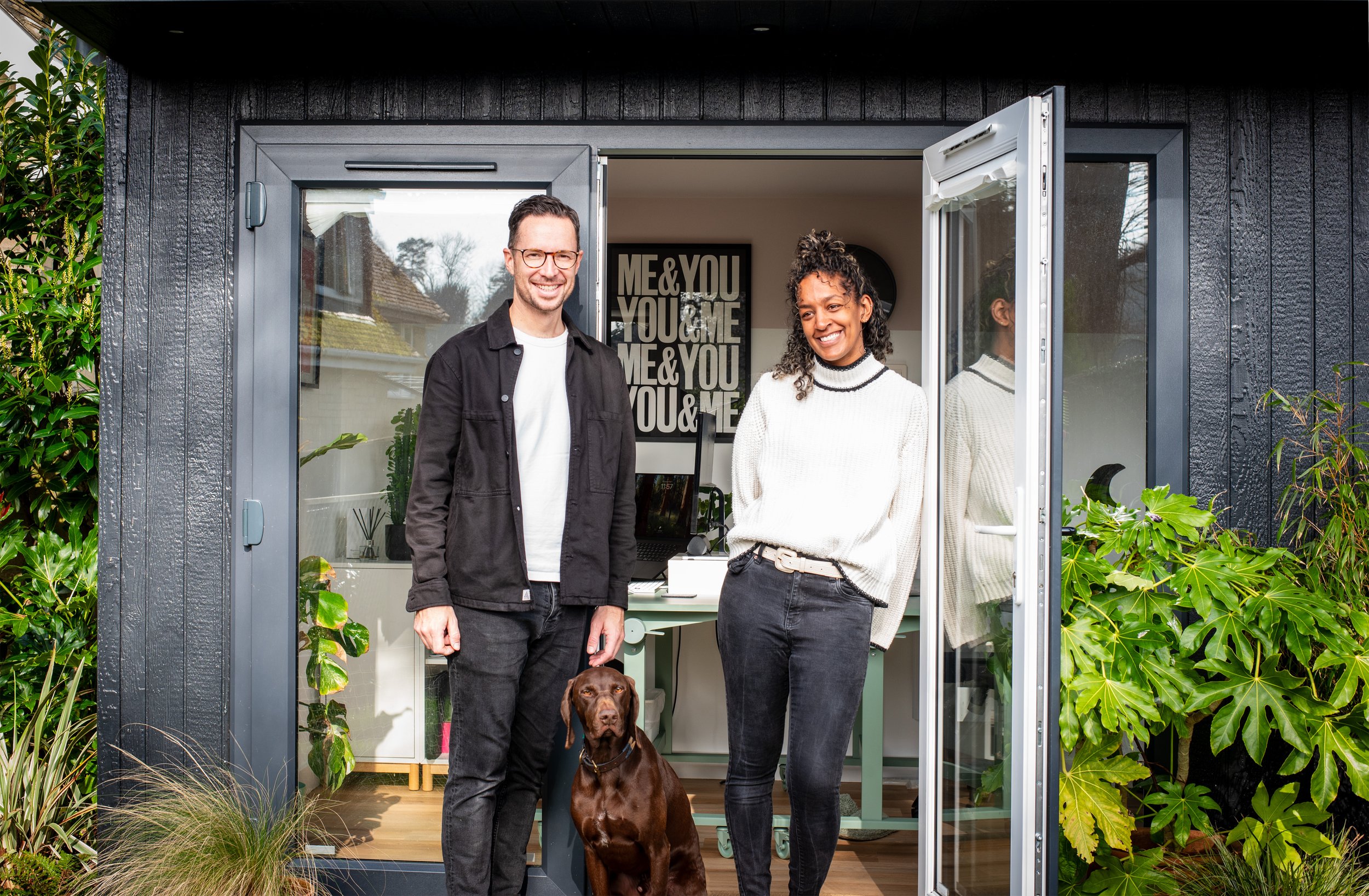Two people and a dog standing in front of an open door to a home. The man is wearing a black jacket and glasses, and the woman is wearing a white sweater. The dog is sitting on the ground between them, and inside the home, there is a framed print with the text 'ME & YOU' repeated multiple times.