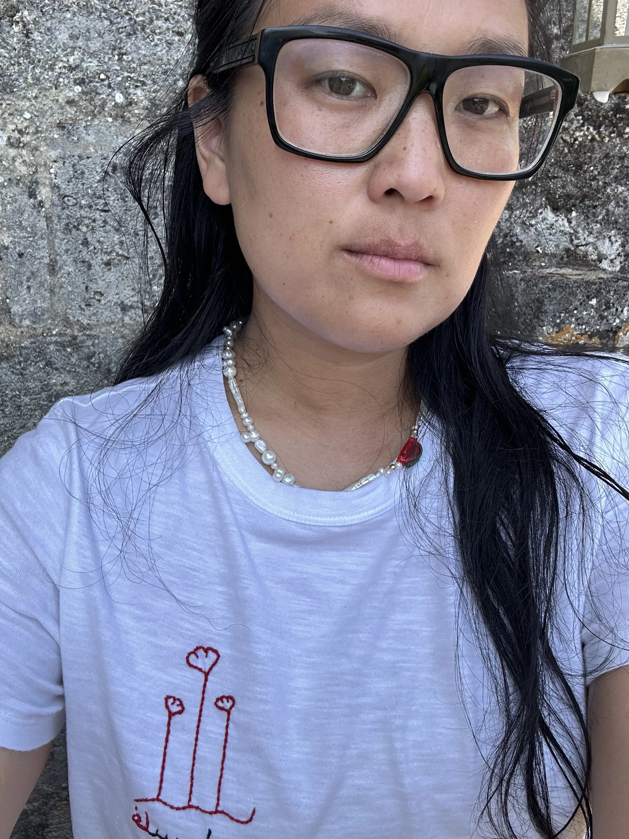 Close-up of a woman wearing large black glasses, a pearl necklace, and a white t-shirt with a red embroidered design, standing outdoors with a stone wall in the background.