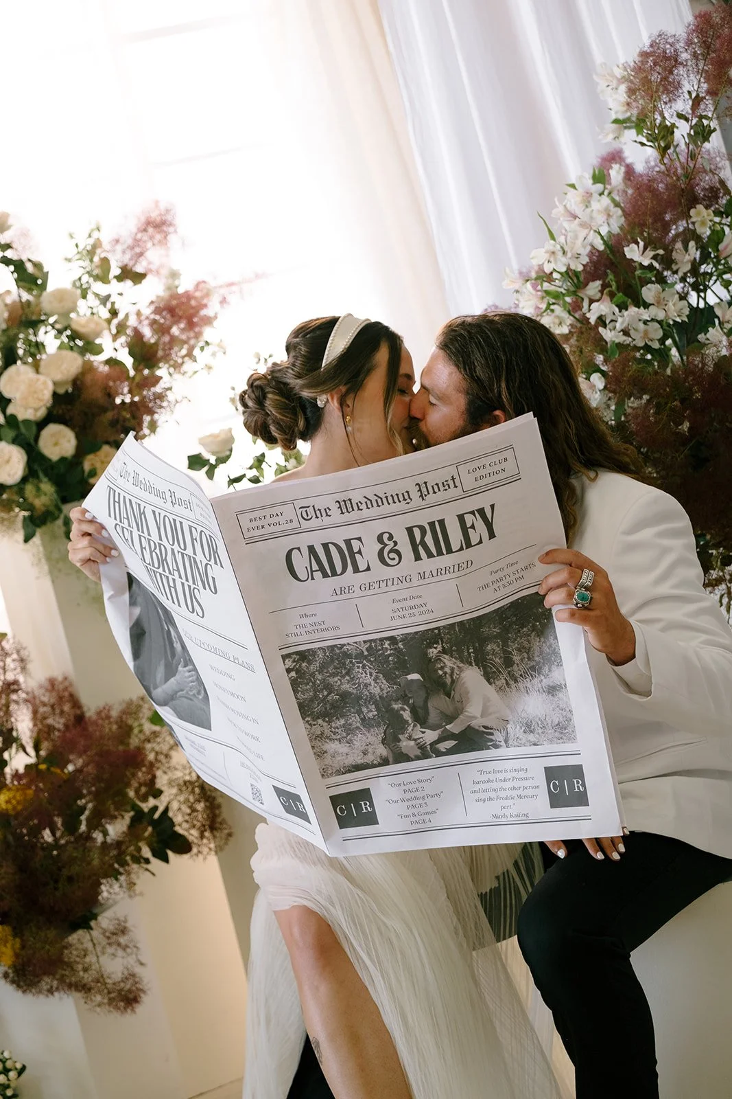 Couple holding a newspaper at their wedding, with the headline 'Cade & Riley are getting married,' surrounded by floral decorations.