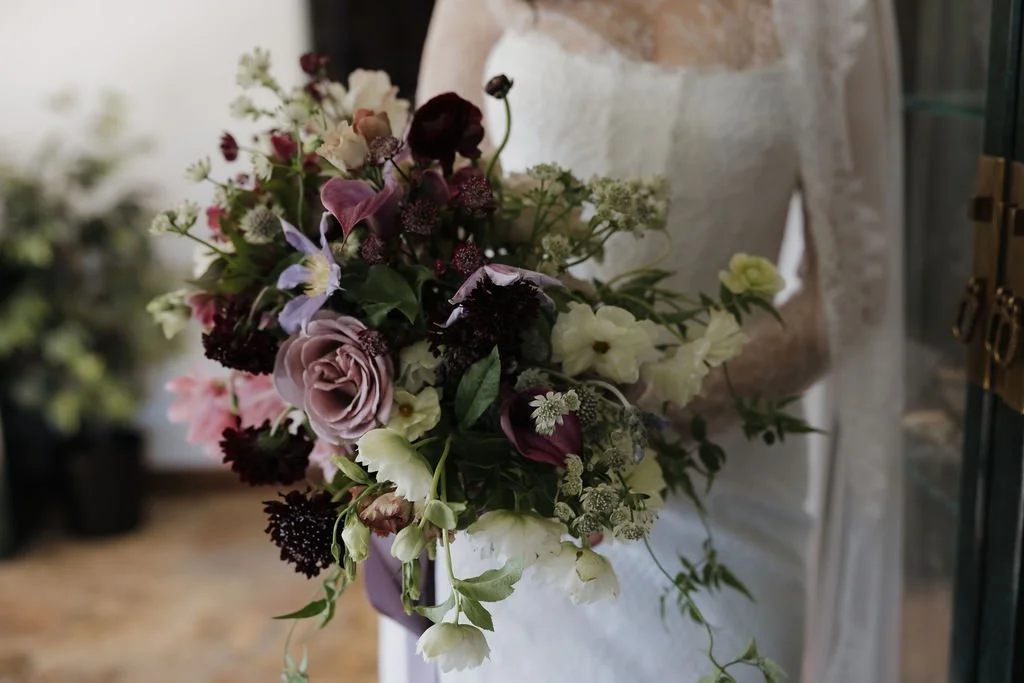 Person holding a bouquet of mixed flowers including roses, calla lilies, and greenery in front of a background with a glass cabinet and potted plants.