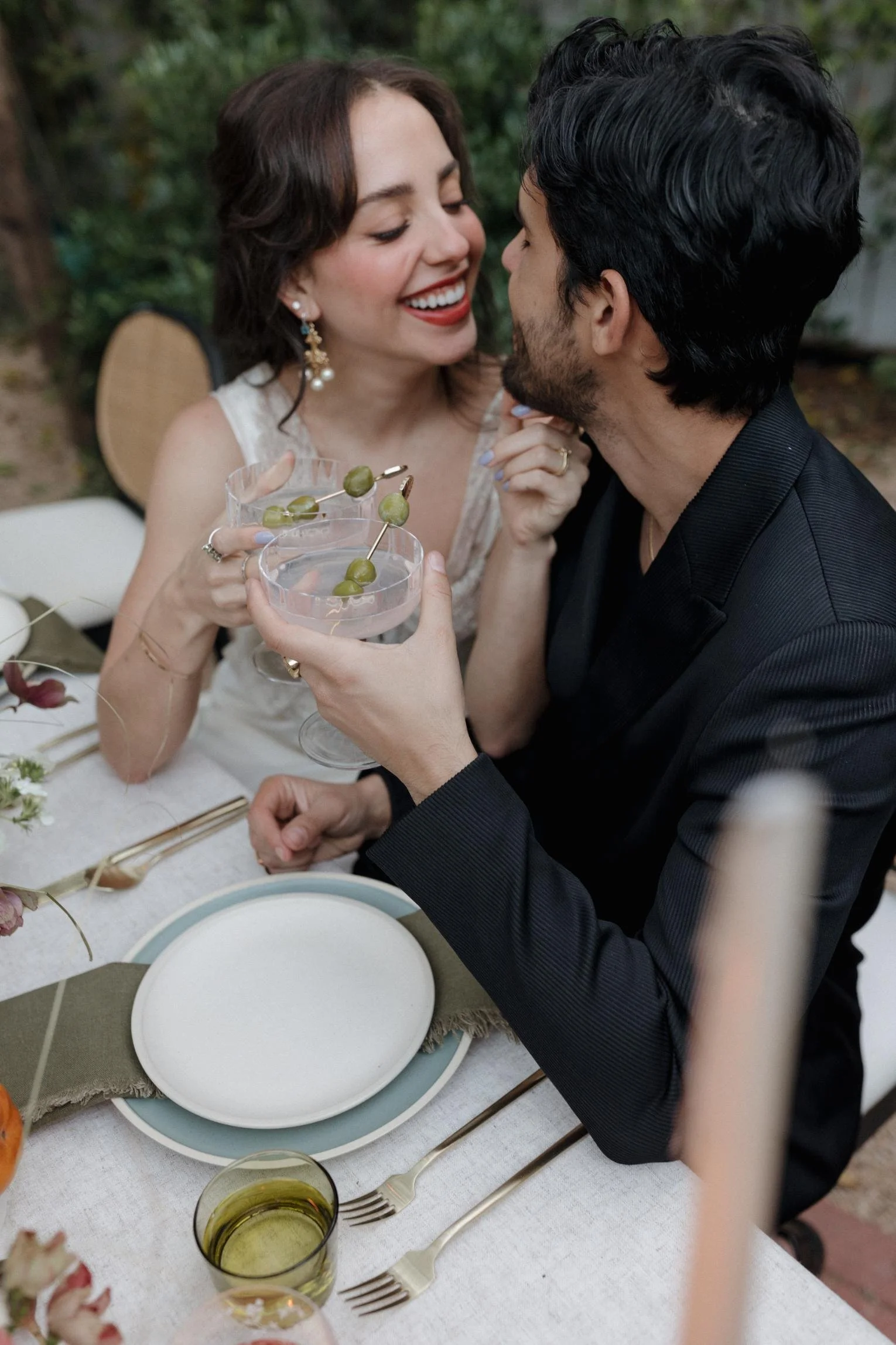A woman and a man are sharing a moment at a dining table, with the woman smiling and leaning towards the man, holding him gently. The man is holding a cocktail glass garnished with green olives. The table is elegantly set with plates, cutlery, and a 