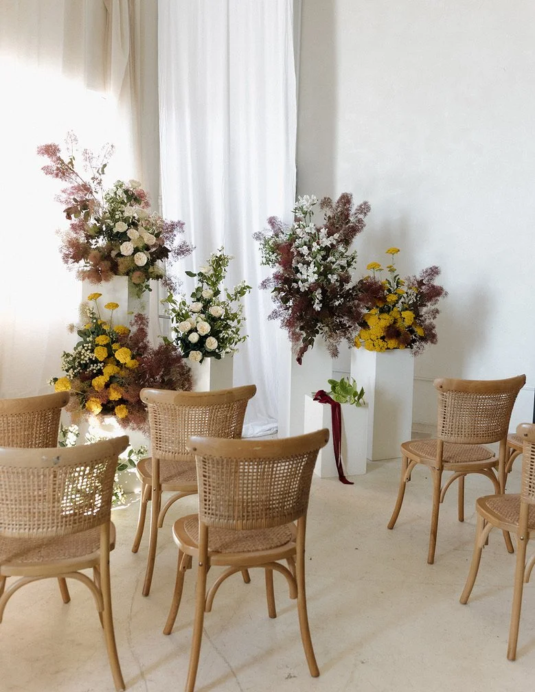 Interior scene with six rattan chairs arranged facing a white wall with large flower arrangements in tall white vases, gathering around a white curtain in a bright room.