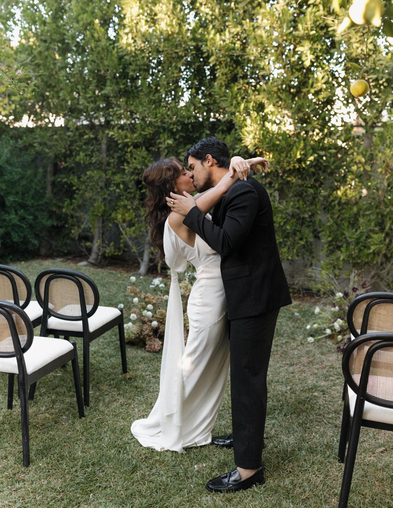 A couple is kissing outdoors in a garden setting, surrounded by green trees and flowers, with chairs in the background.