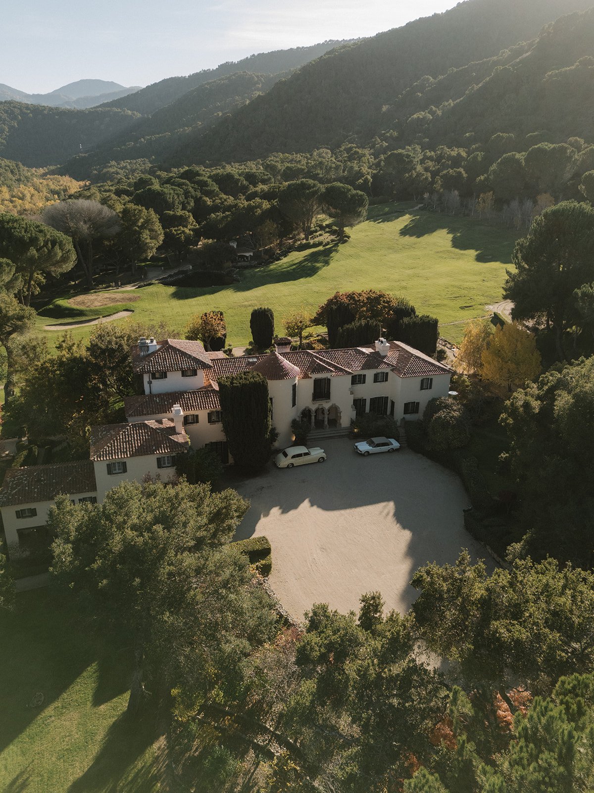 A large white mansion with a red-tiled roof, parked cars, and surrounding lush greenery, set in a mountainous landscape.