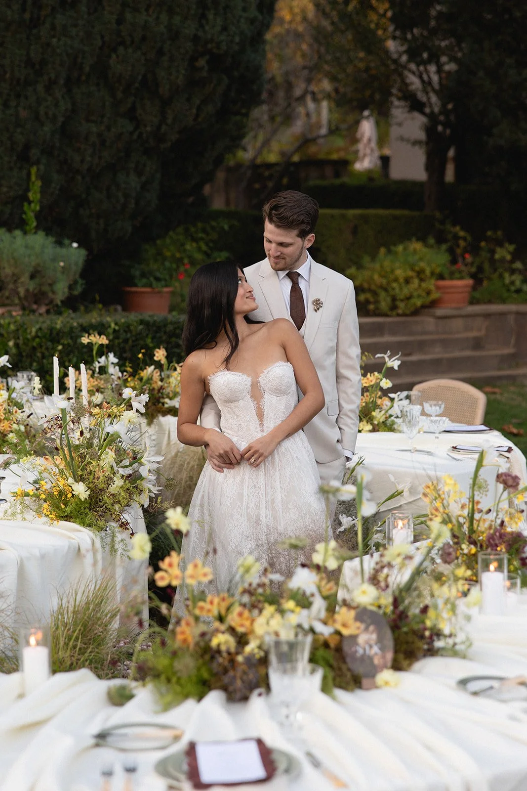 A bride and groom standing together outdoors at a wedding reception, surrounded by floral arrangements and decorated tables.