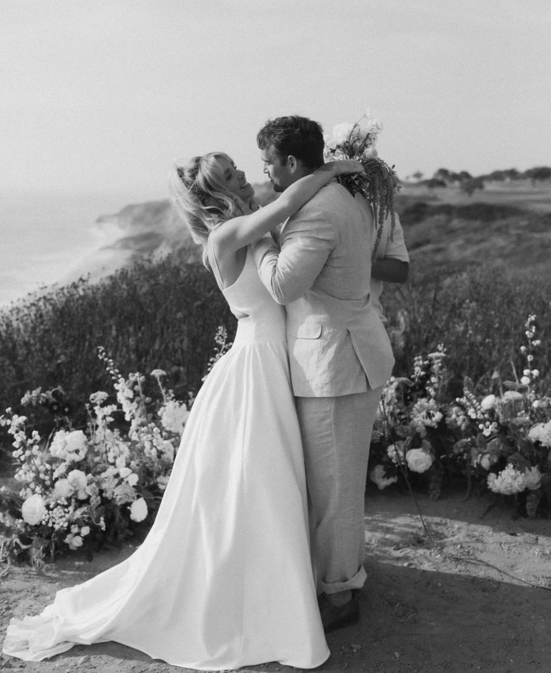 A couple in wedding attire embracing on a beach with flowers in the background, black and white photo.
