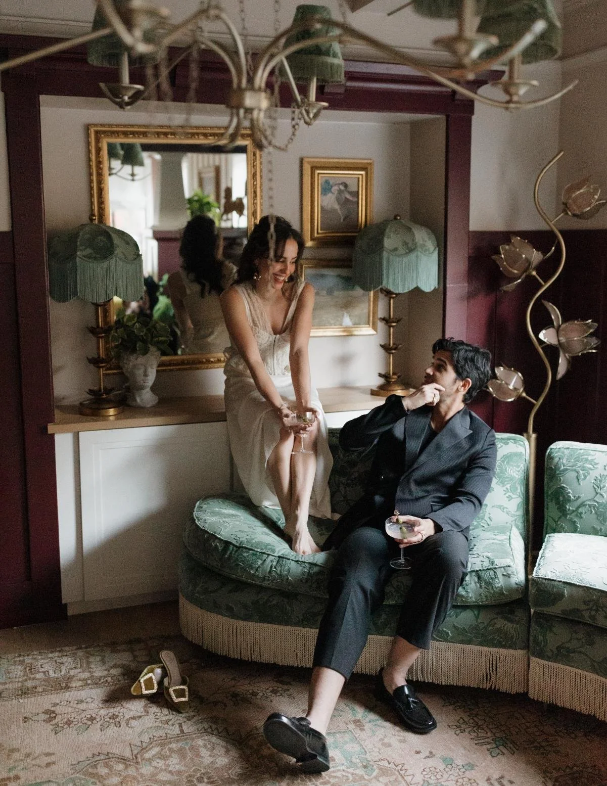 A woman in a white dress sitting on a cabinet, leaning over a couch, laughing, while a man in a dark suit, sitting on the couch, drinks a cocktail and looks at her. The room has green floral upholstery, a large mirror, framed art, and vintage lamps.