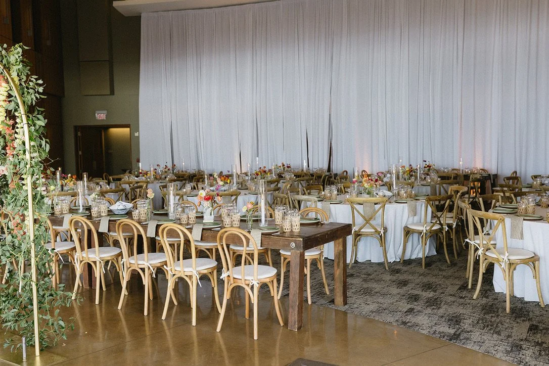 Elegant banquet hall with multiple round tables decorated with floral centerpieces, candles, and tableware, set for a formal event with wooden chairs around the tables and long white curtains in the background.