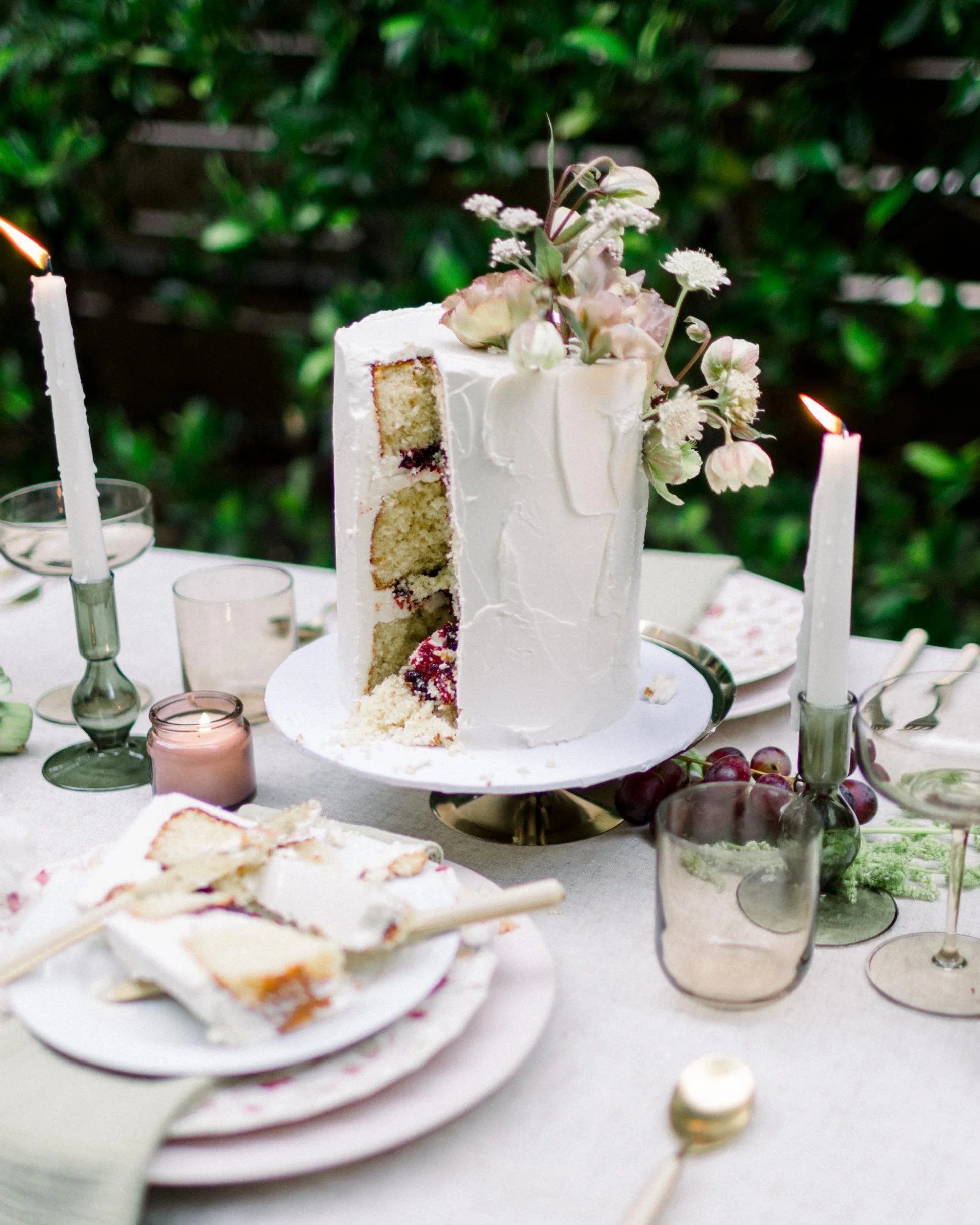 Decorative white birthday cake with floral arrangement on top, partially sliced to show layers of sponge cake with jam filling, on a table with candles, plates, and glasses outdoor setting.