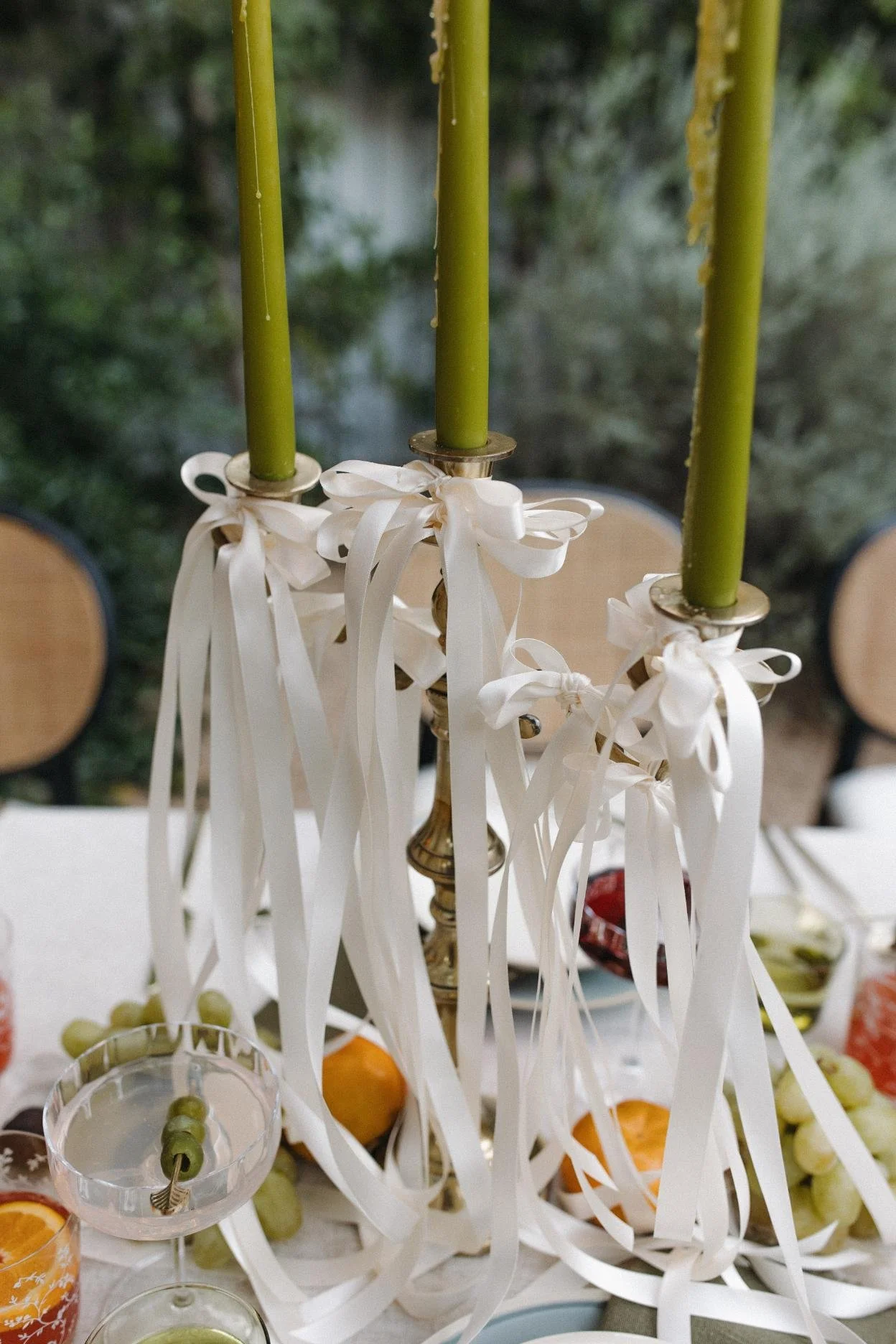 A chandelier with three green candles tied with white ribbons, with a background of a dining table set with fruit and drinks and chairs.
