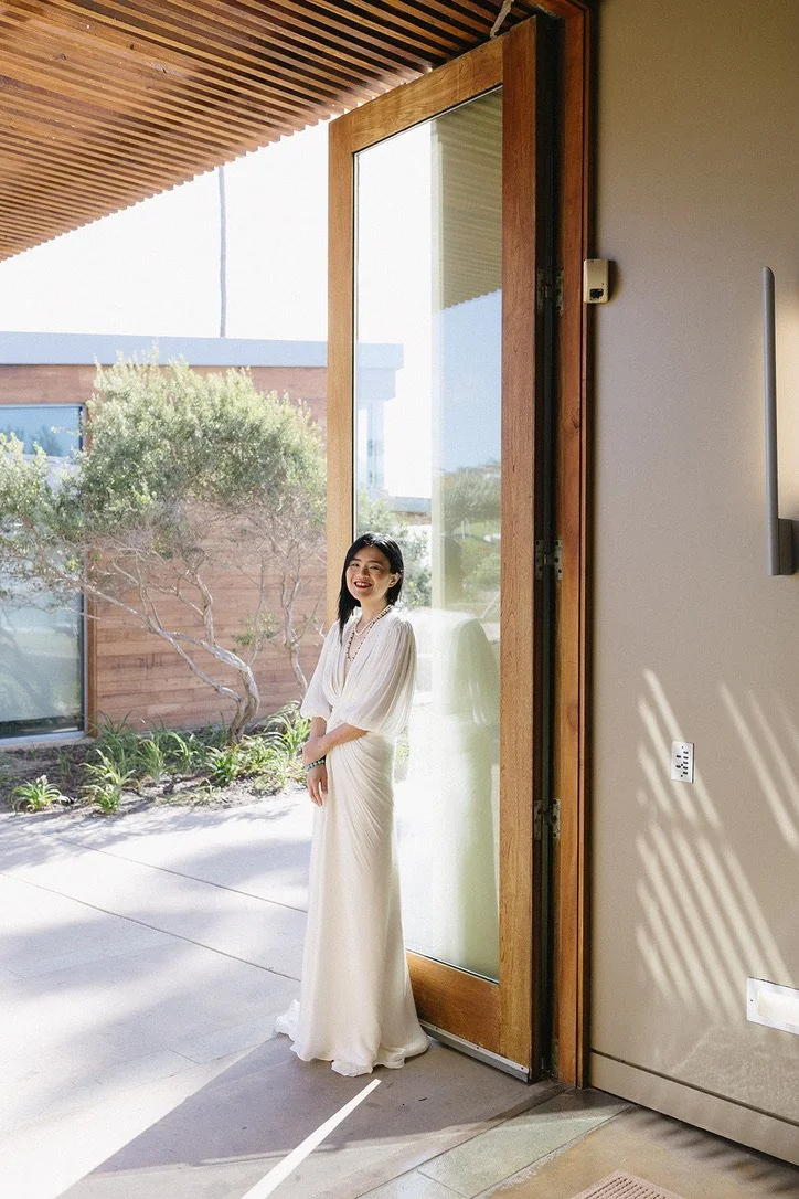 A woman in a white dress standing in a doorway with a view of a garden outside.